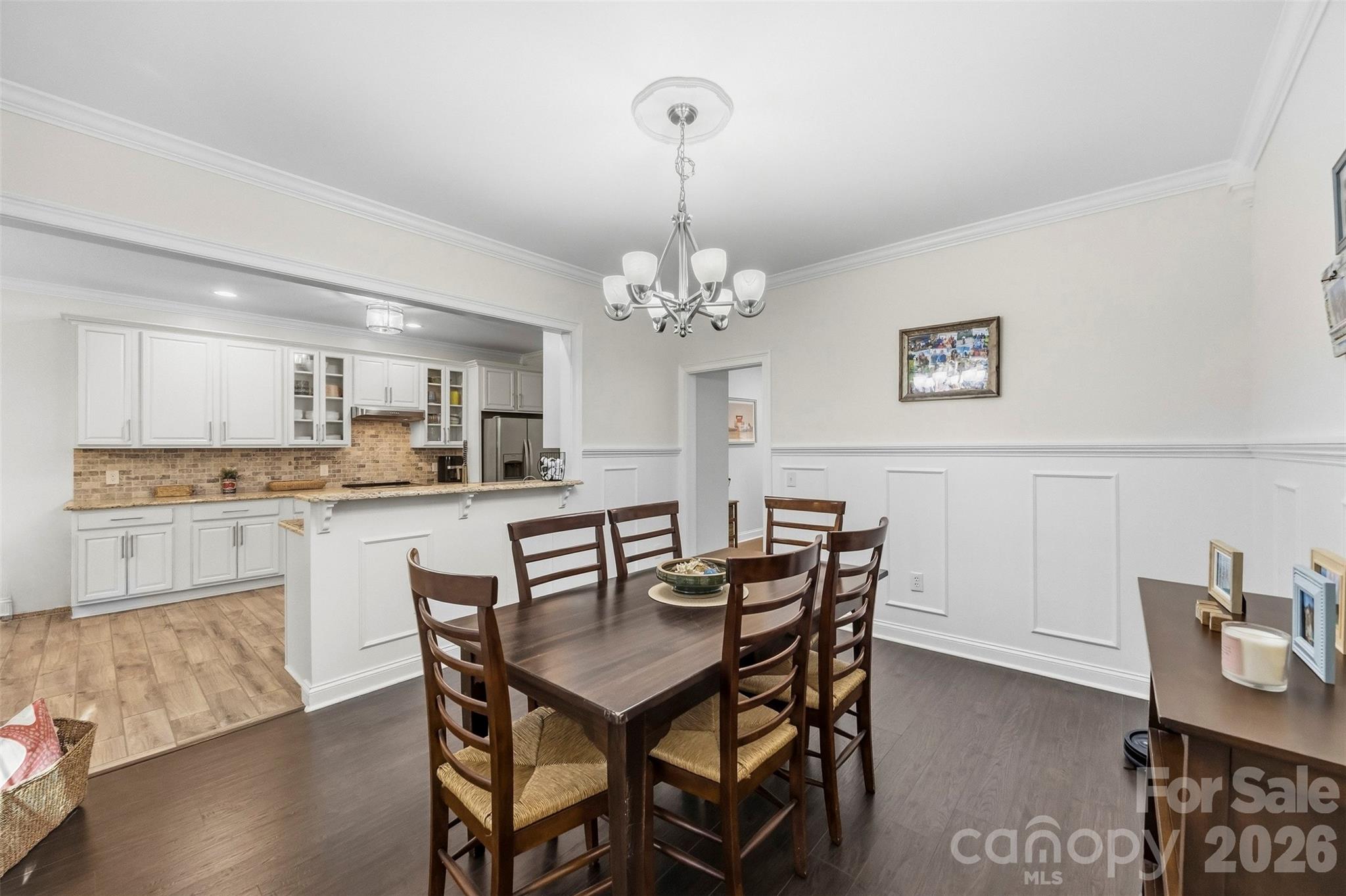 Dining room with dark wood table and chairs beside an open kitchen with white cabinets and brick backsplash.