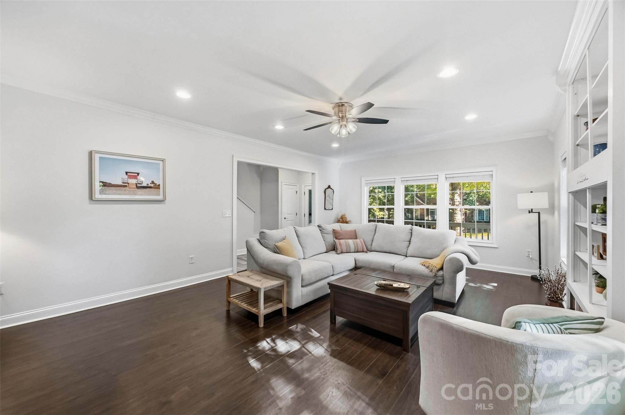 Furnished living room with a gray sectional, dark wood floors, built-in shelves, and three large windows.