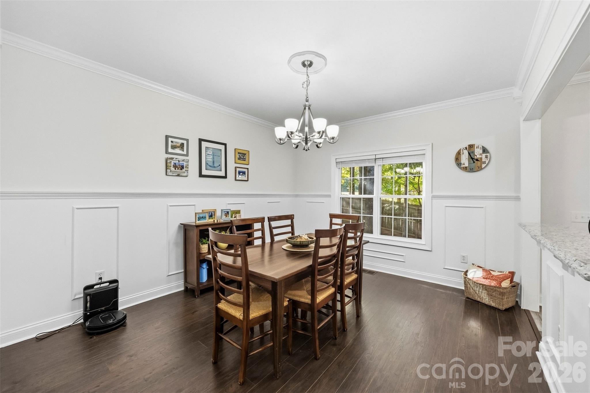 Dining room with dark wood table, white paneled walls, chandelier, and a large window.