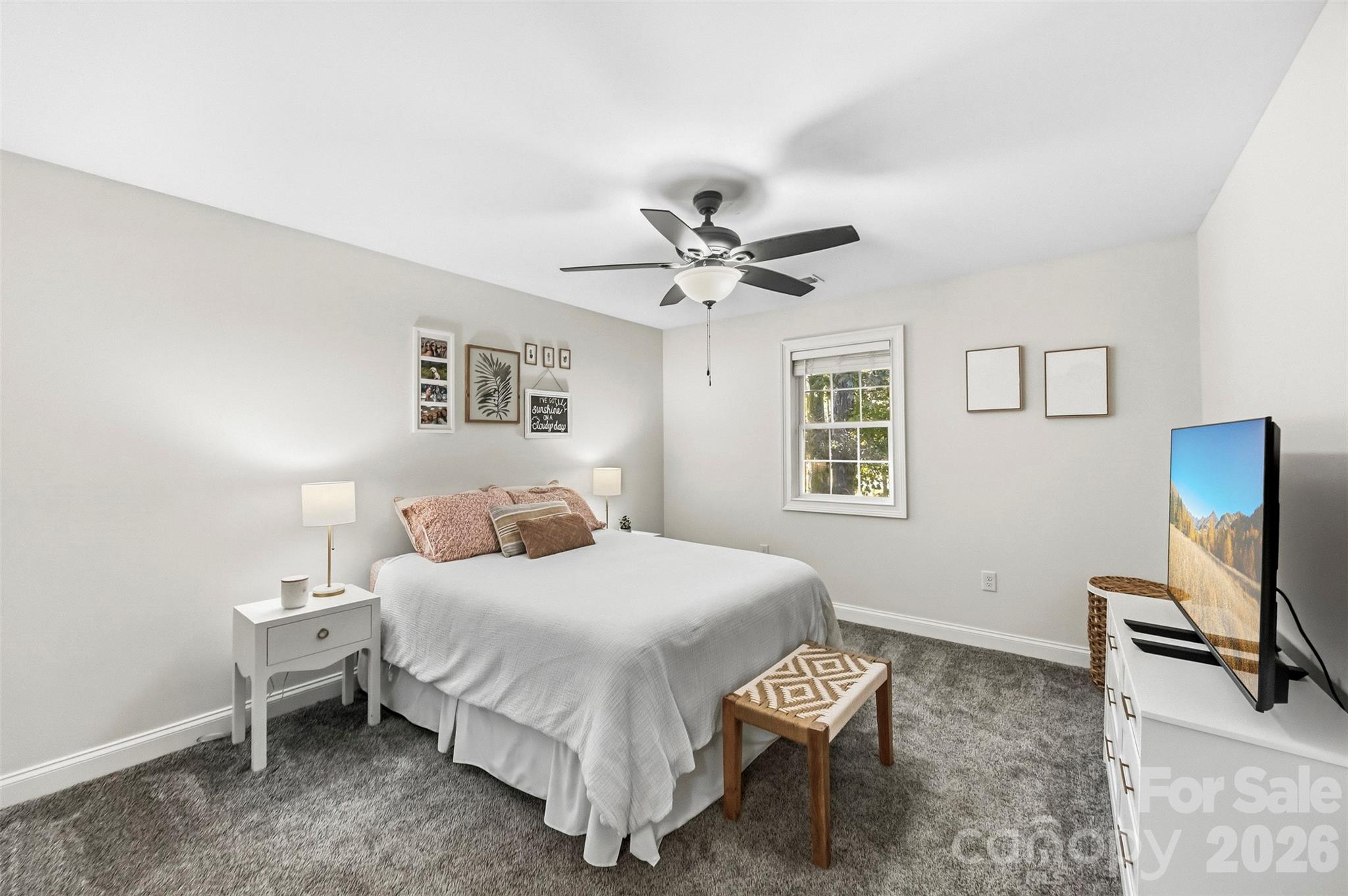Furnished bedroom with a white bed, gray carpet, ceiling fan, and television.