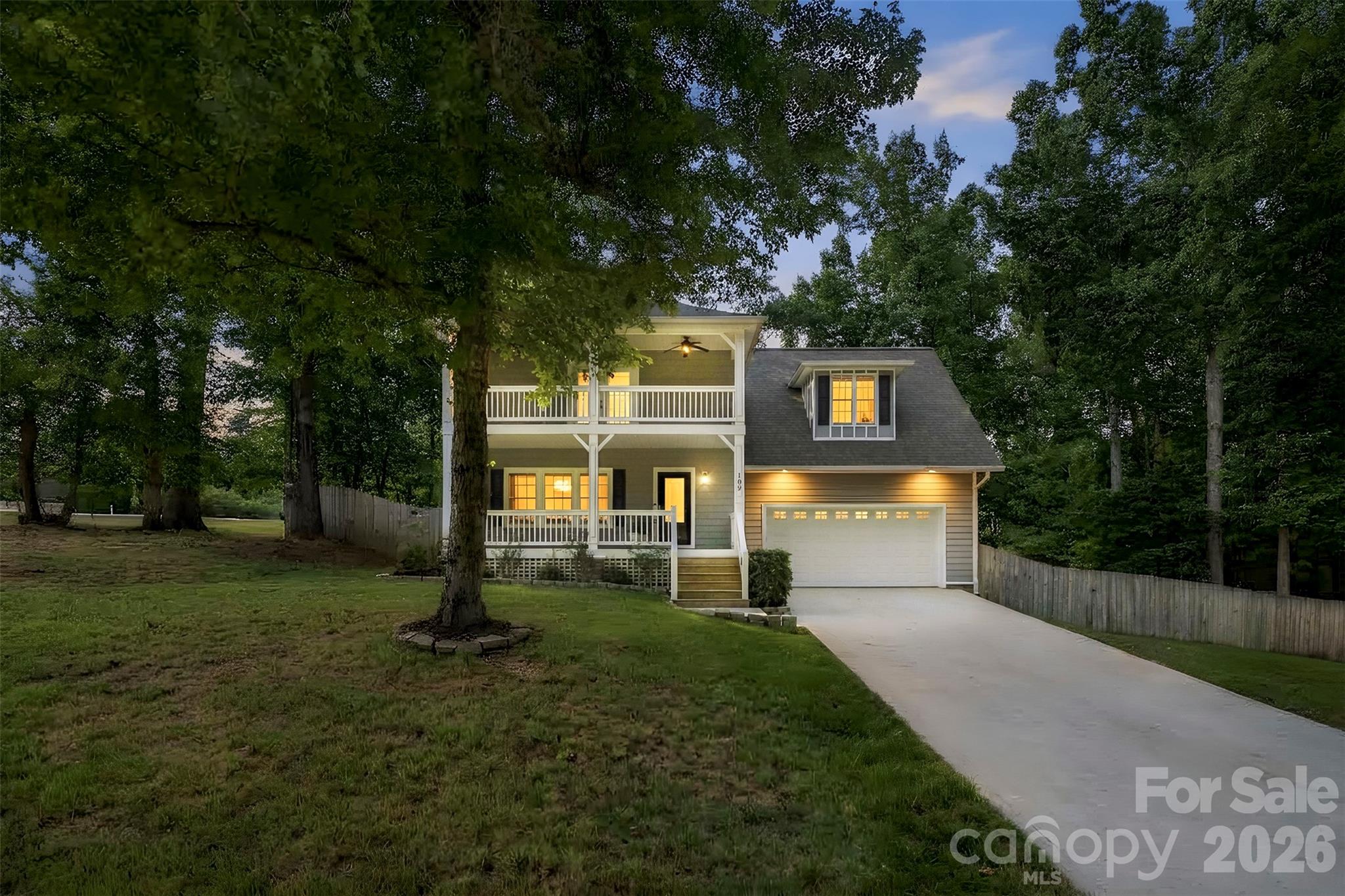 Two-story gray home with white porches and an attached garage, warmly lit at dusk among mature trees.