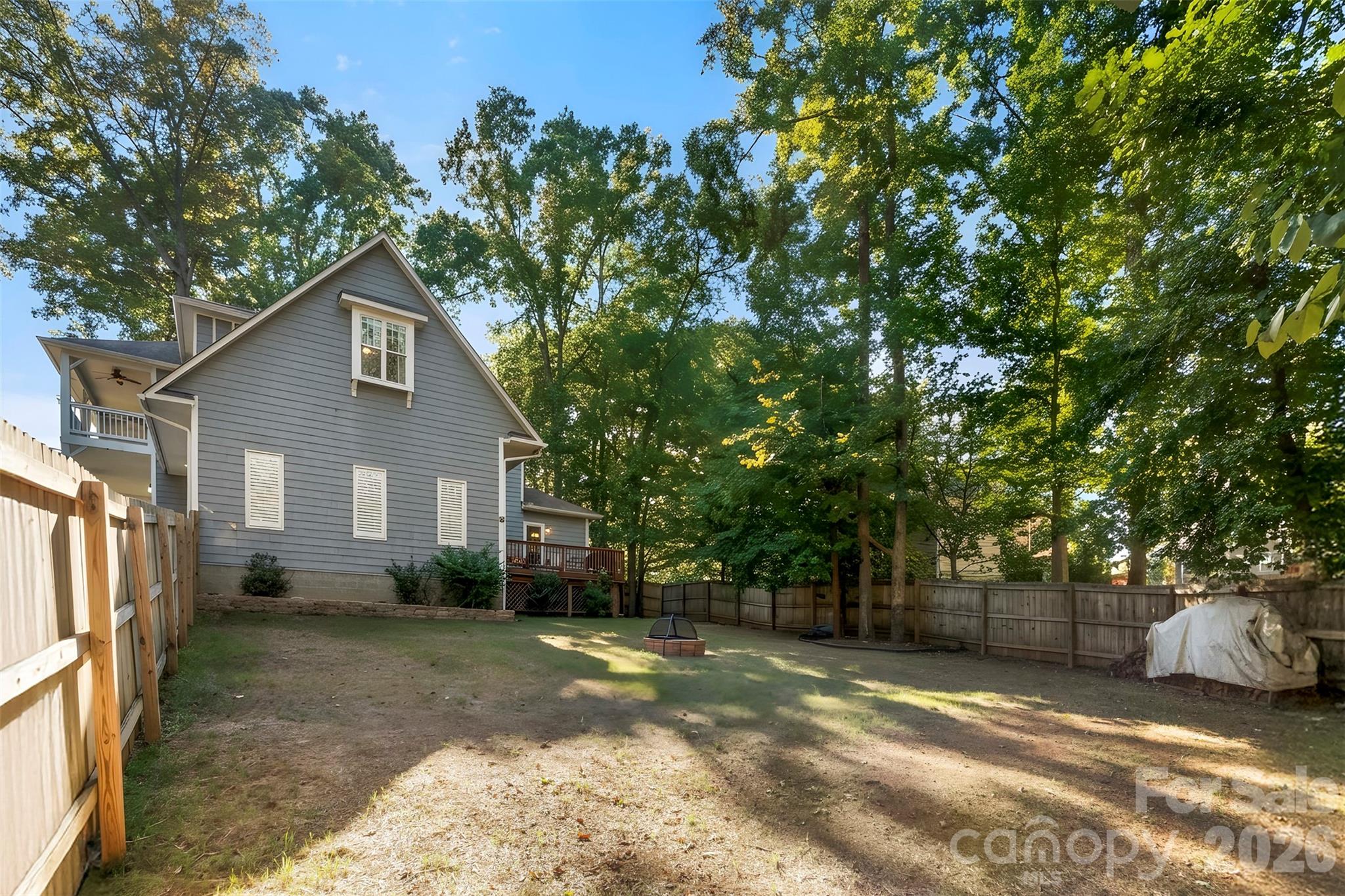 Gray-sided house beside a fenced, tree-shaded backyard with a fire pit and raised deck.