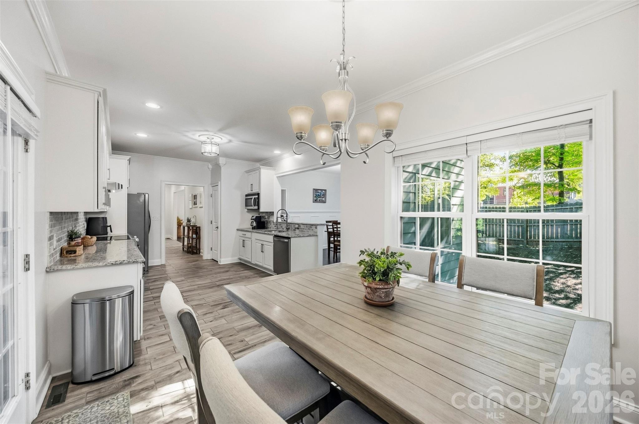 Open kitchen and dining area with a long table beside large windows overlooking a fenced yard.