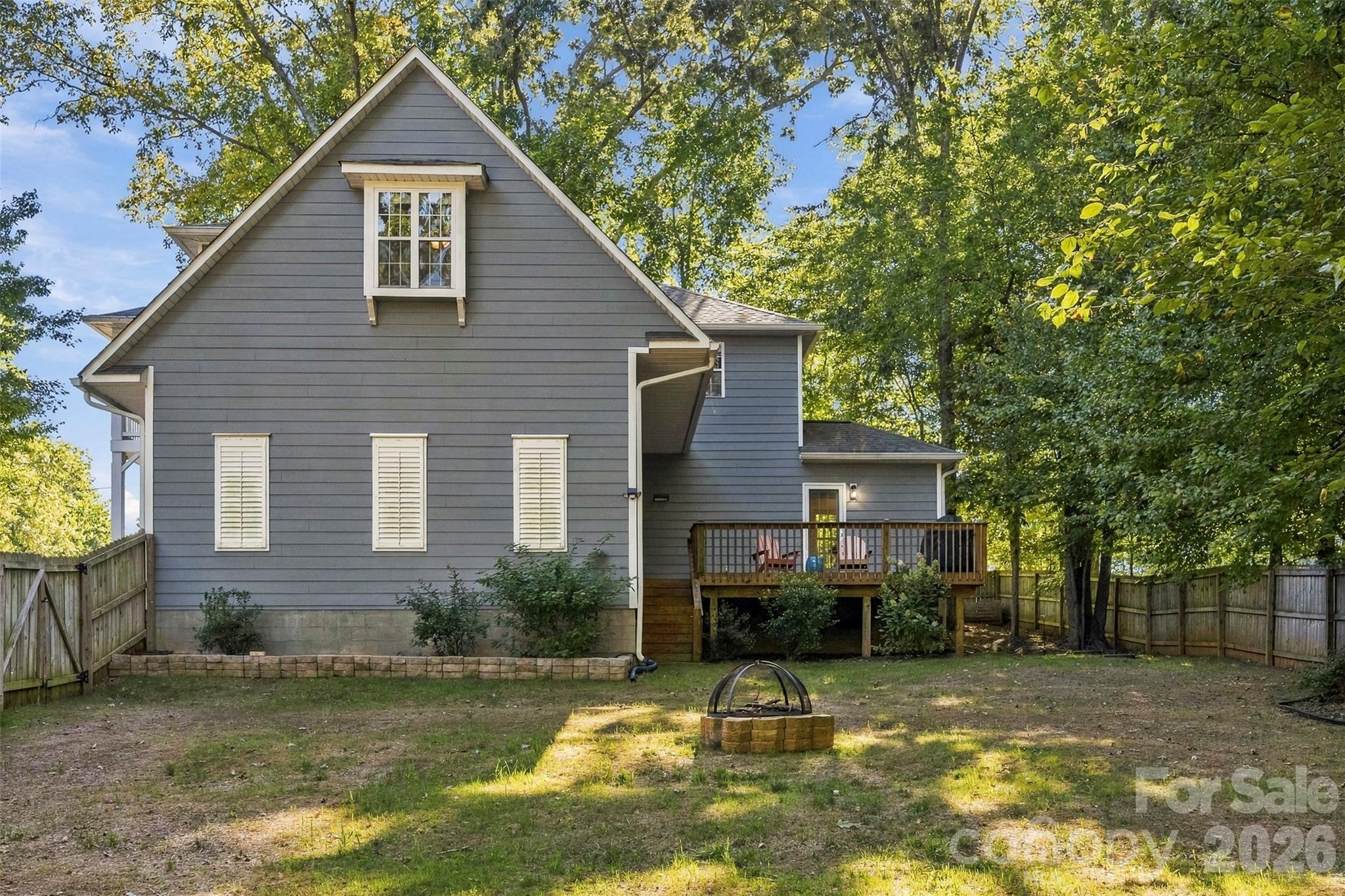 Gray gabled house with a raised deck, fenced yard, and stone fire pit beneath mature trees.