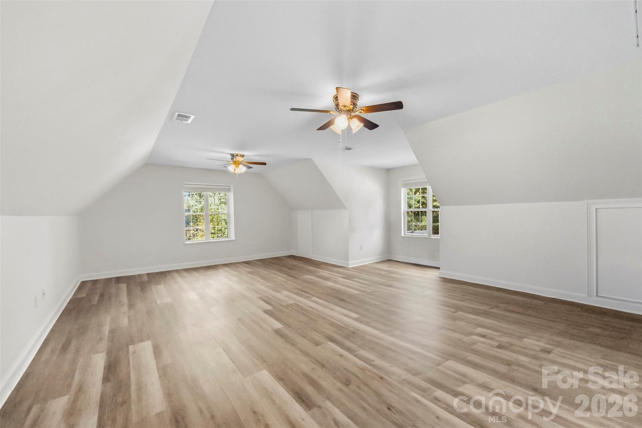 Finished upper-level room with sloped ceilings, wood-look flooring, two ceiling fans, and windows overlooking greenery.