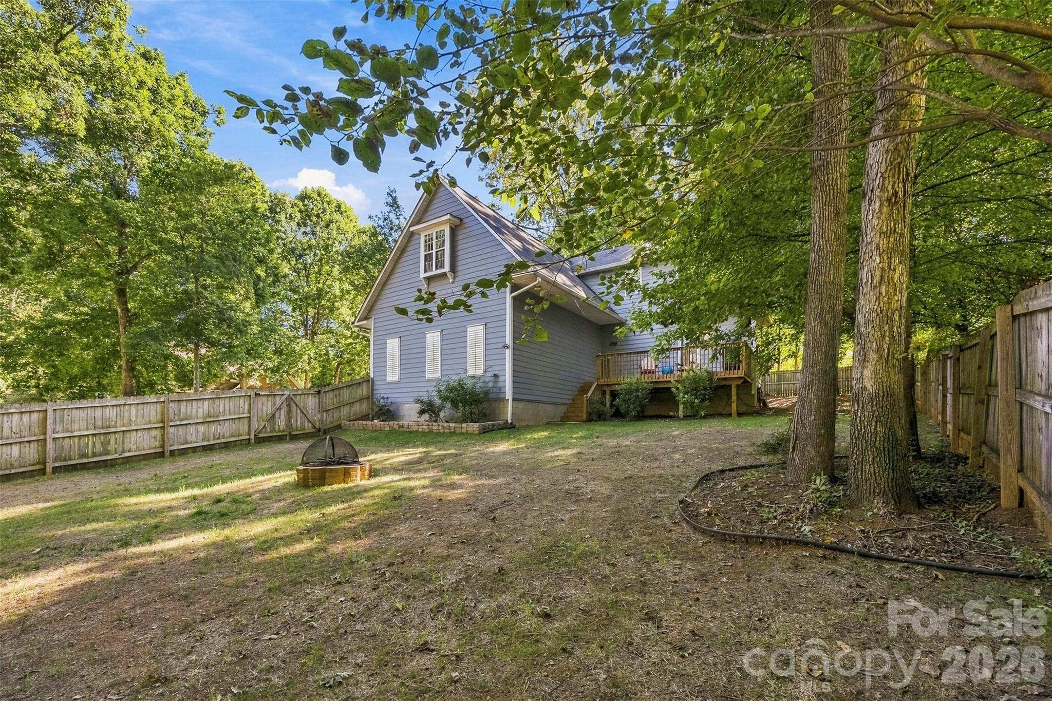 Gray two-story house behind a fenced, tree-shaded backyard with a fire pit and raised deck.