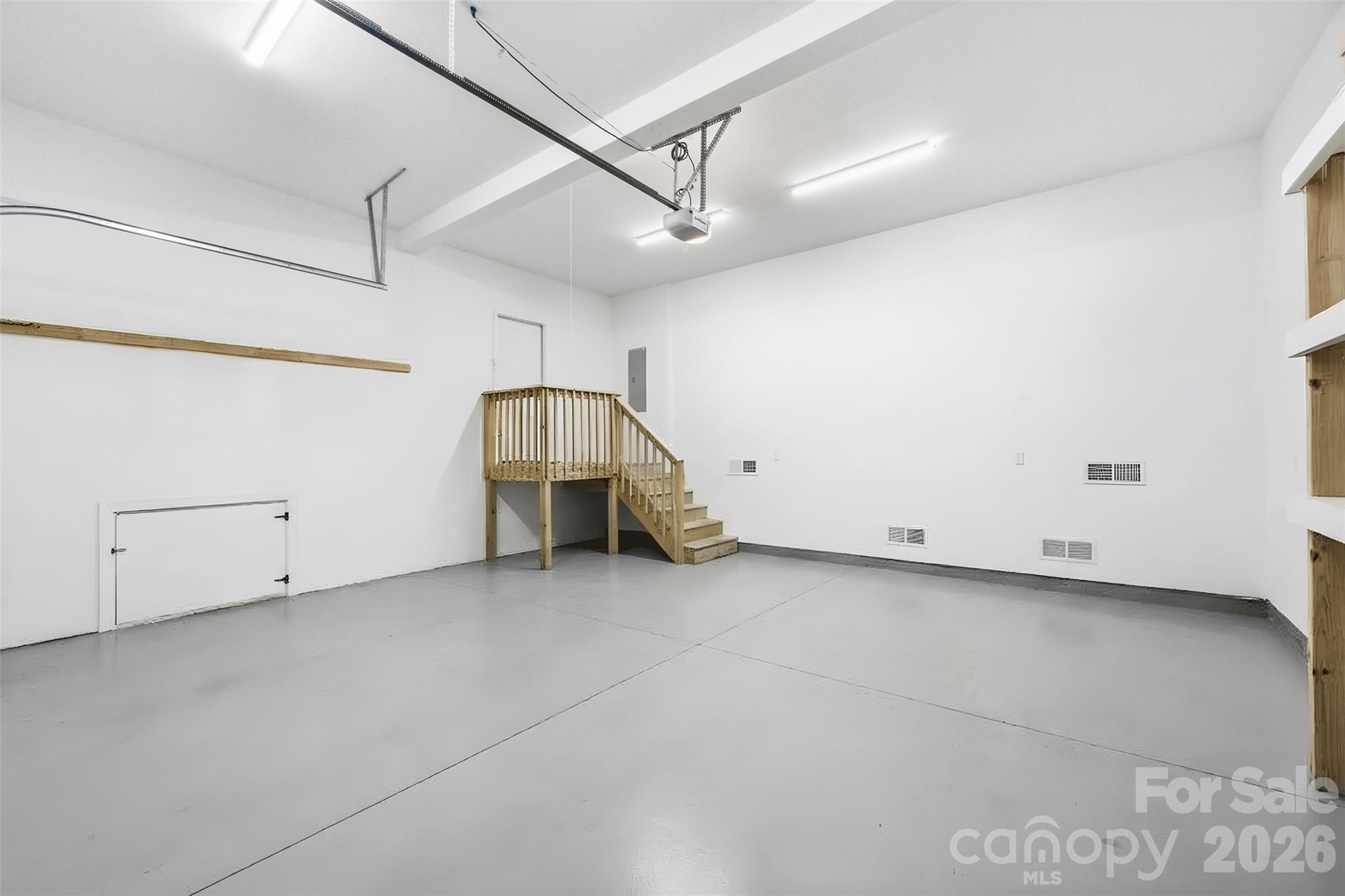 Vacant garage with gray floor, overhead door tracks, and wooden stairs to a raised platform.