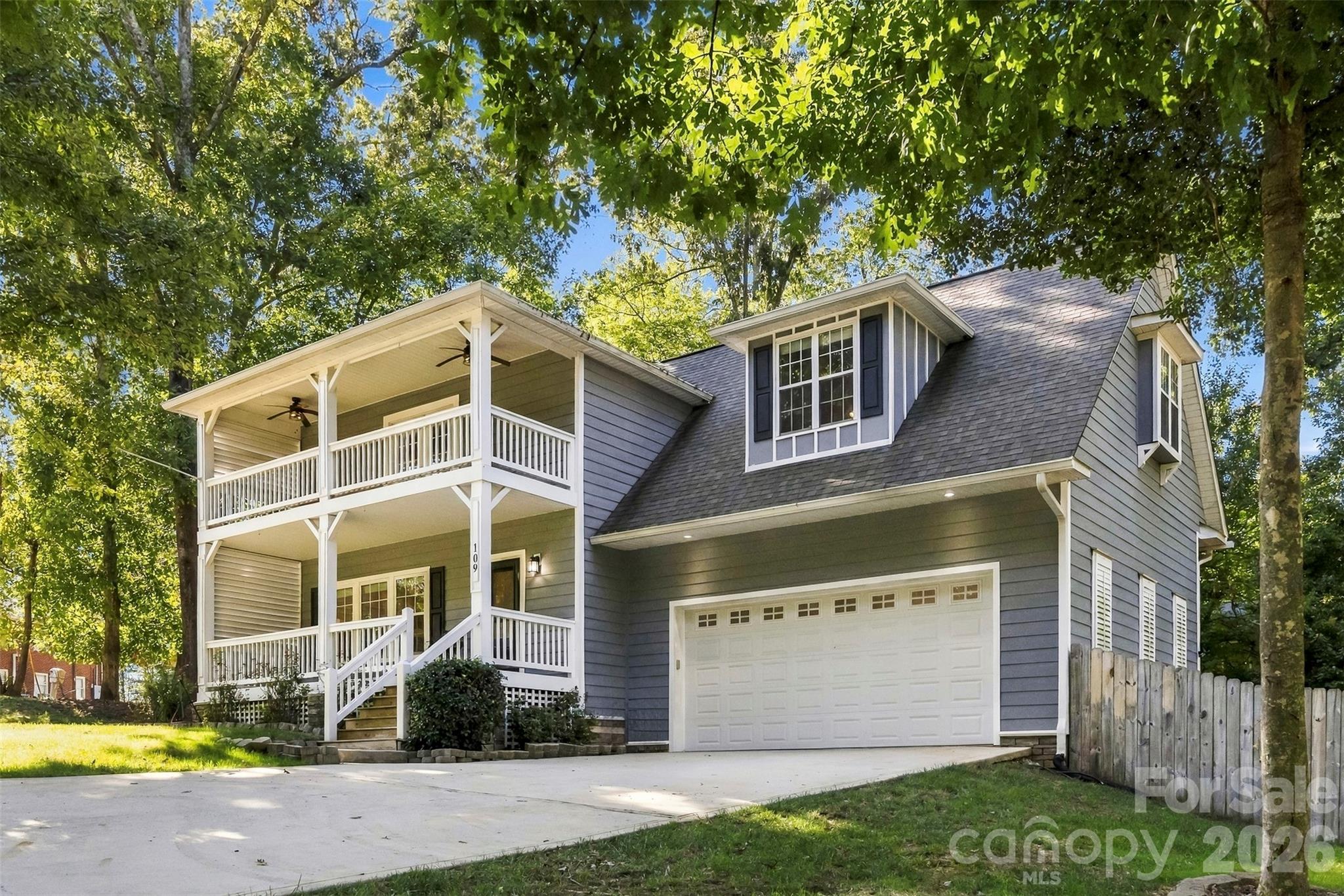 Gray two-story home with a covered two-level porch, balcony, and attached garage beneath mature trees.