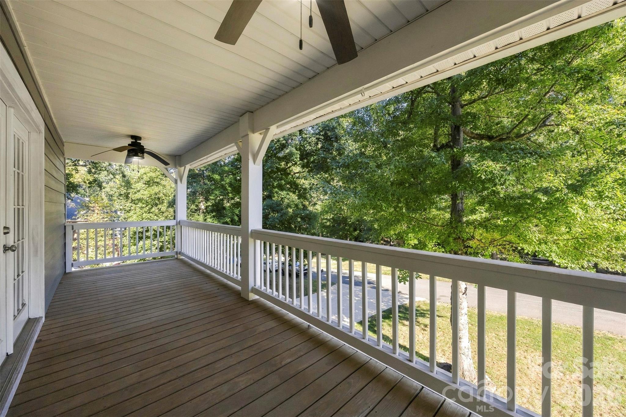 Covered porch with brown flooring, white railings, ceiling fans, and leafy trees beyond.
