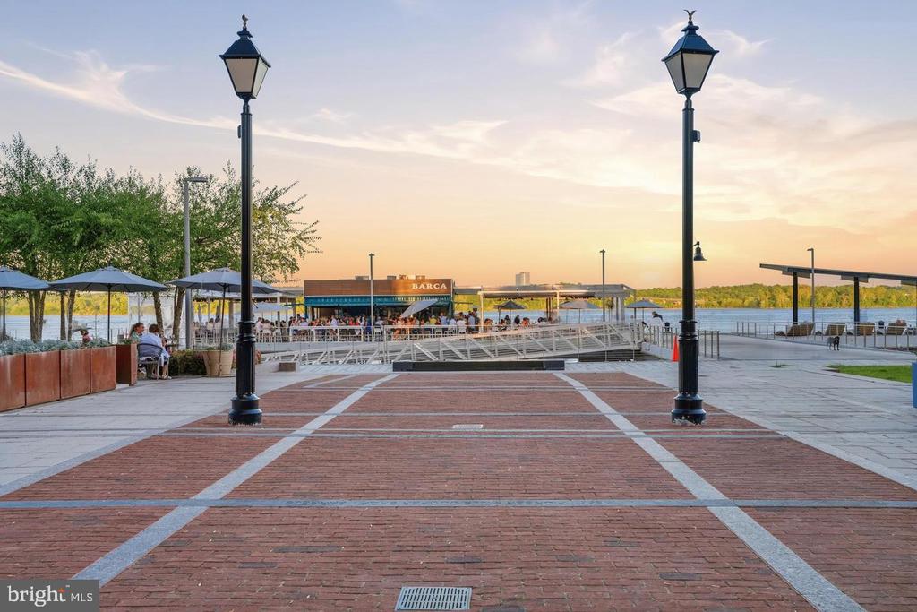 Waterfront promenade with brick paving, lampposts, outdoor dining, and the BARCA restaurant beside the river.
