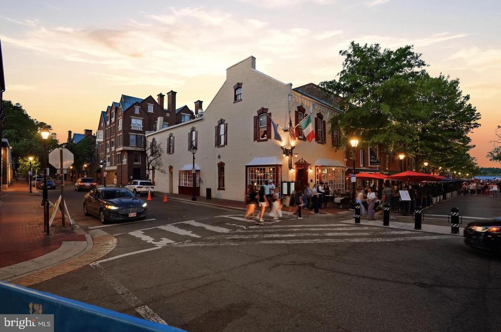 Alexandria street corner at dusk with a white building, restaurant patio, and pedestrians crossing.