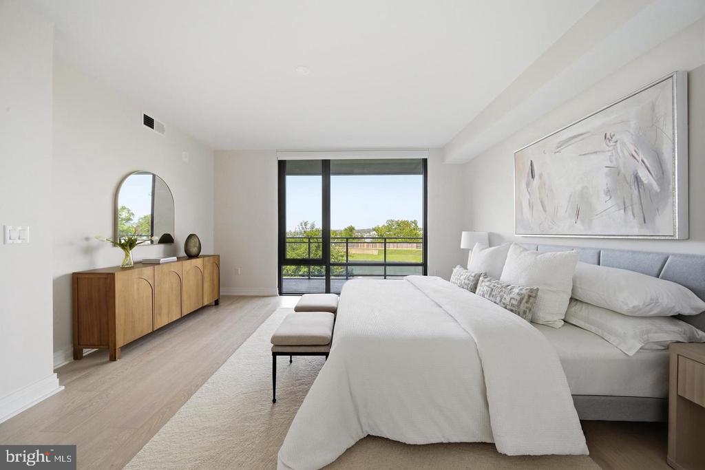 Staged bedroom with a large white bed and sliding glass doors overlooking green lawn and trees.