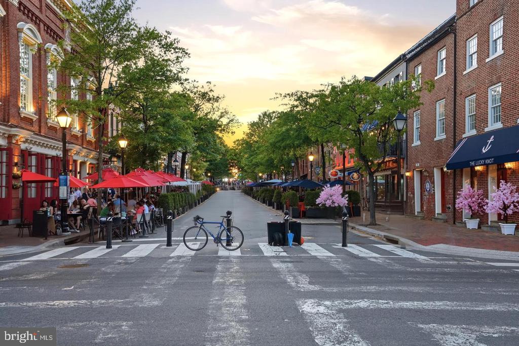 Tree-lined street with brick storefronts, outdoor dining, and red umbrellas at golden hour.