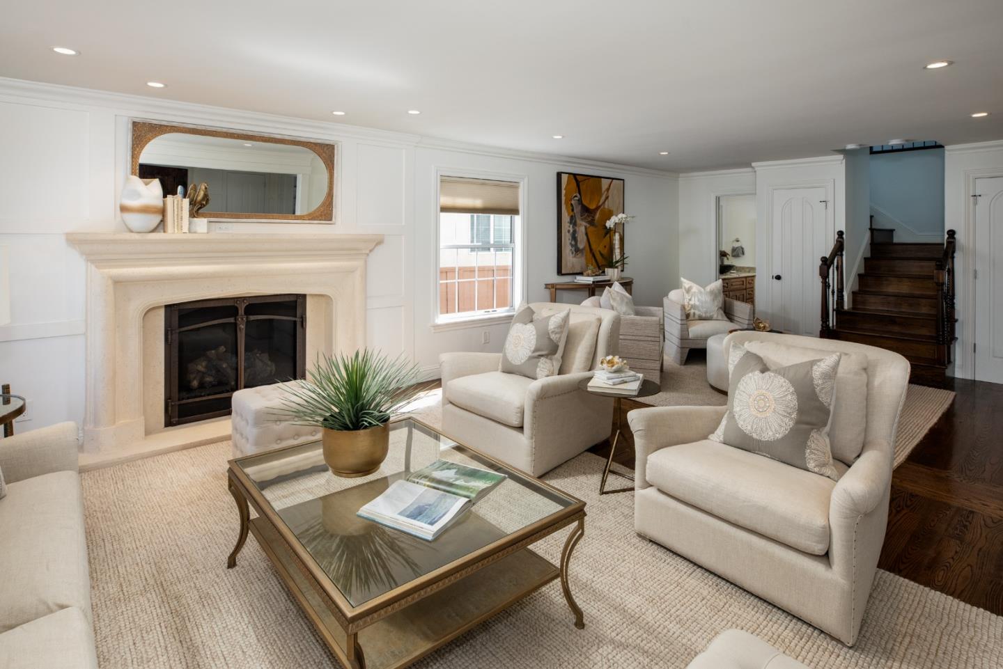 Staged living room with a cream fireplace, light upholstered seating, and a dark wood staircase.