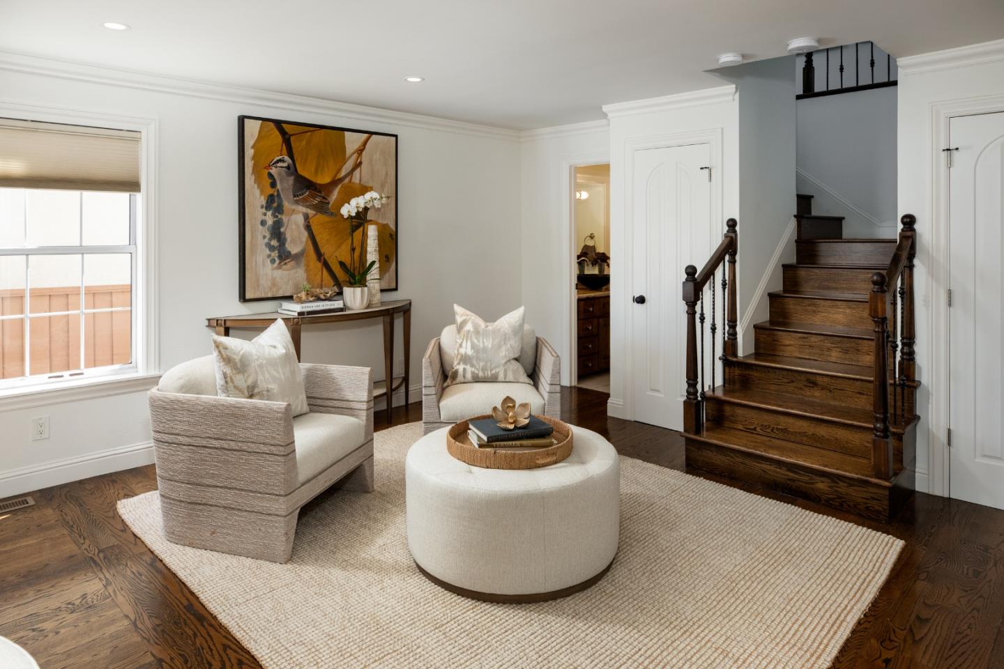 Living room with cream armchairs, a round ottoman, and a dark wood staircase.