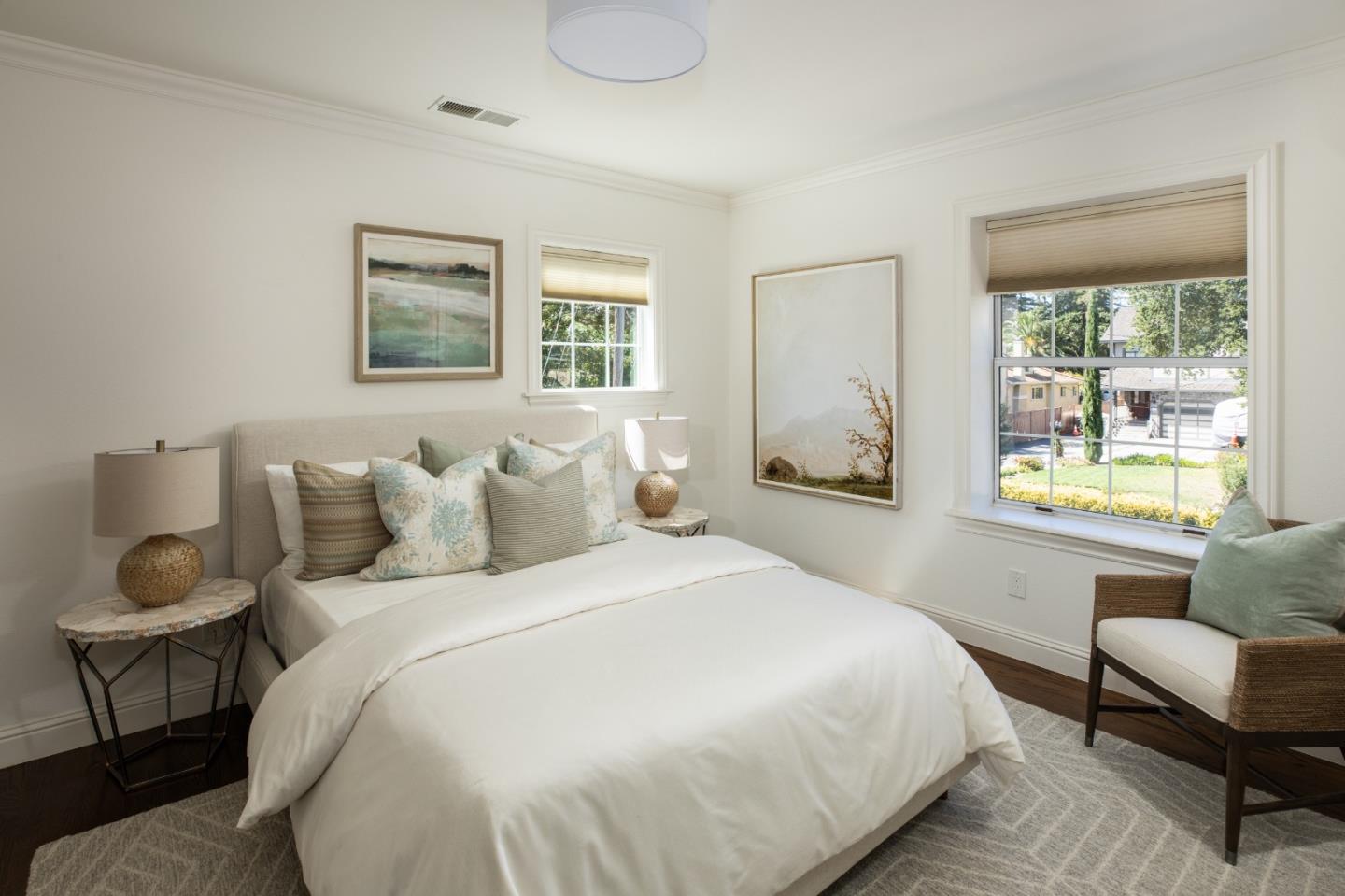 Staged bedroom with a white bed, decorative pillows, and windows overlooking the street.