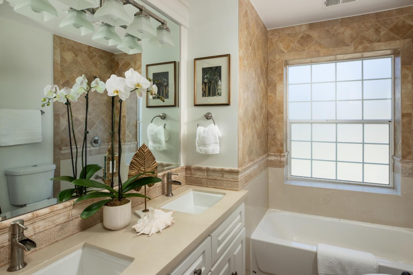 Bathroom with a double-sink vanity, tan stone tile, and a bathtub beneath a large gridded window.