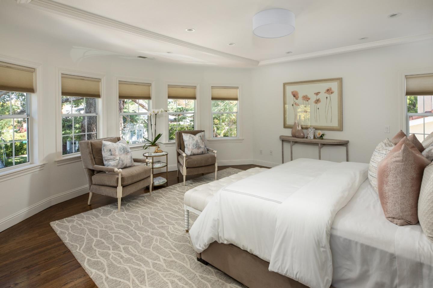 Staged bedroom with a white bed, two armchairs, and multiple windows with shades.