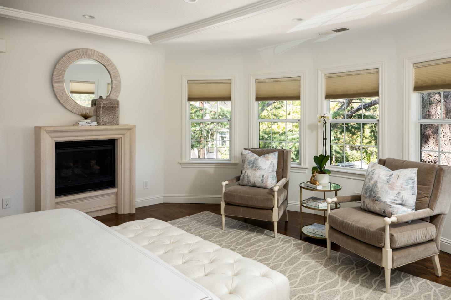 Bedroom with a fireplace, two upholstered chairs, and tall windows overlooking leafy trees.