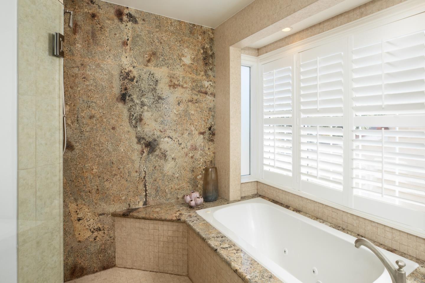 Bathroom soaking tub with mottled stone surround, white plantation shutters, and glass shower partition.