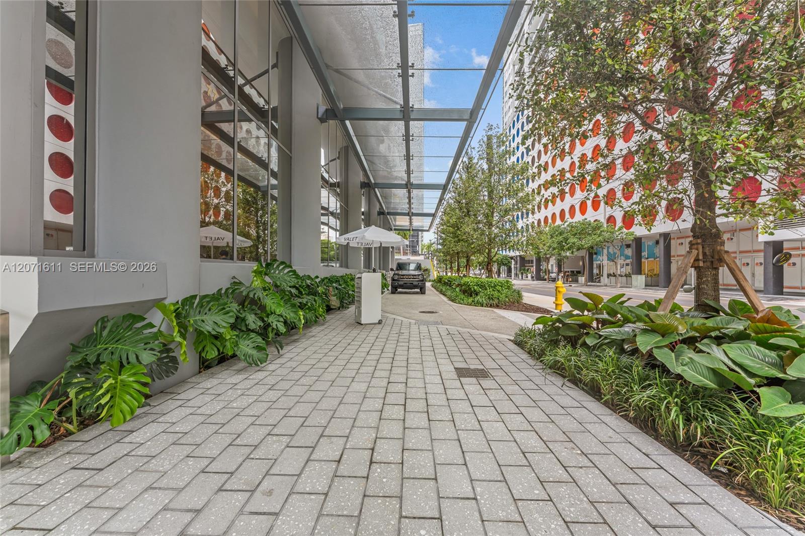 Modern building walkway with a glass canopy, tropical landscaping, and a colorful circular-patterned facade across the street.