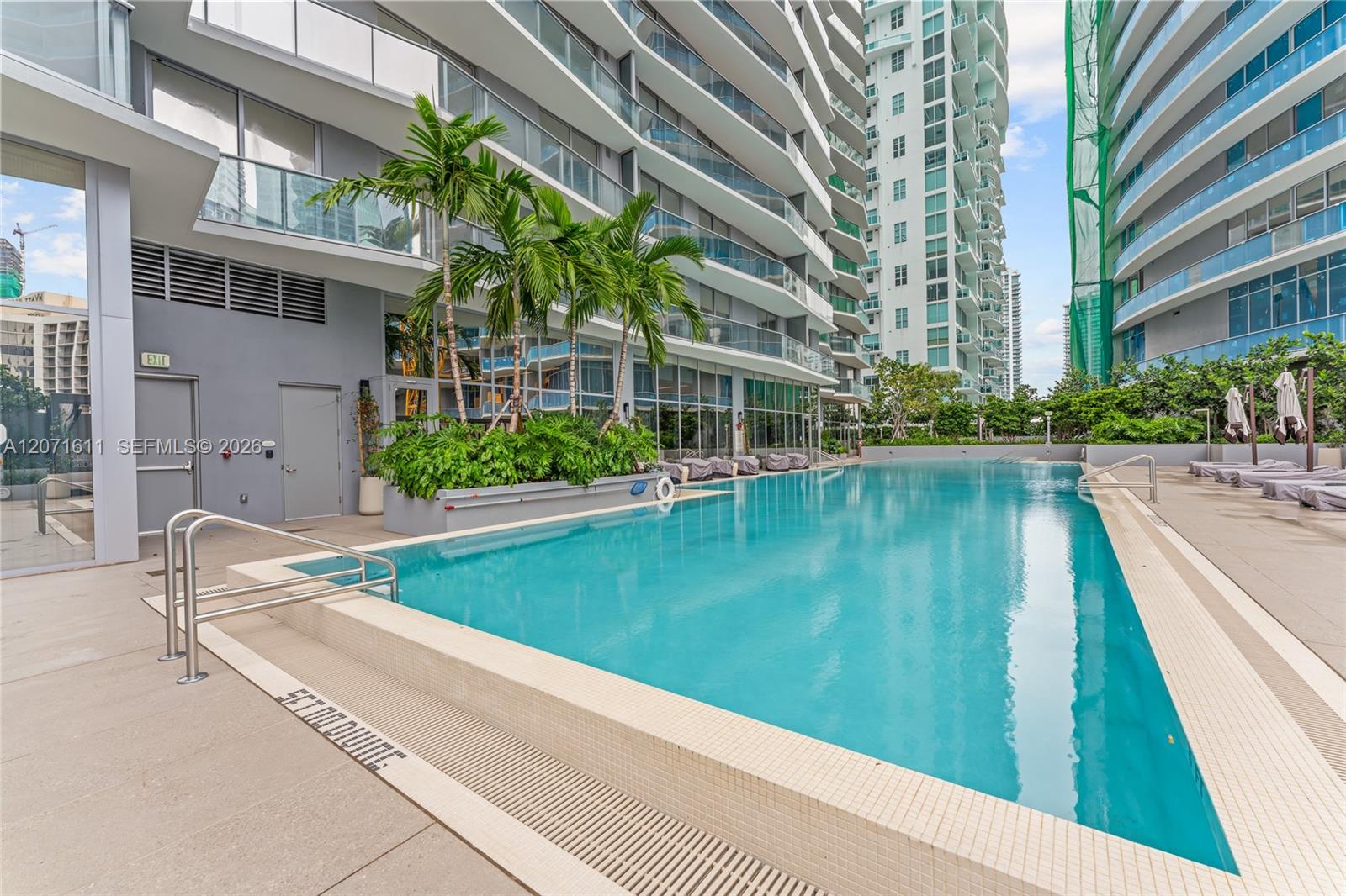 Outdoor condominium pool between high-rise buildings, with palm trees and lounge chairs.