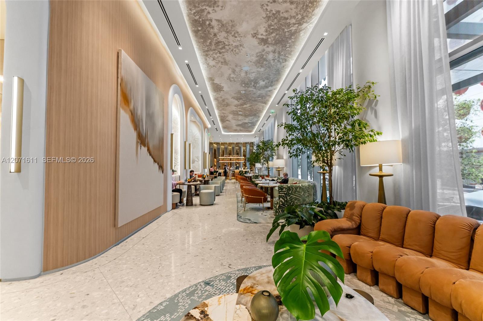 Furnished lobby lounge with orange leather seating, indoor trees, and a long marble-floored corridor.