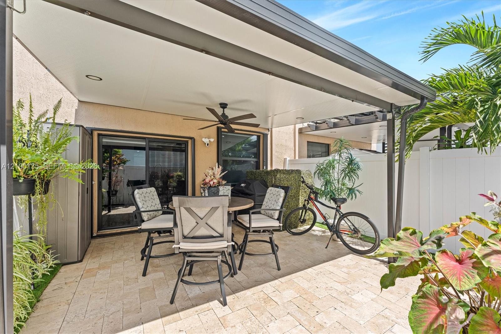 Covered patio with a stone-paver floor, outdoor dining set, ceiling fan, and potted plants.