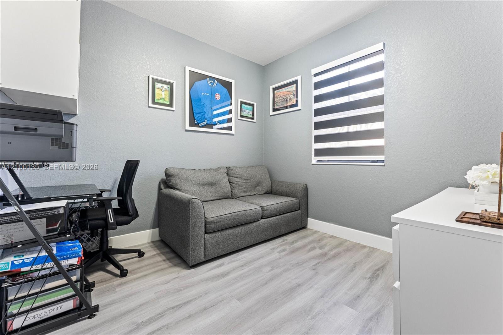 Home office with a gray loveseat, desk, framed sports memorabilia, and striped window blinds.