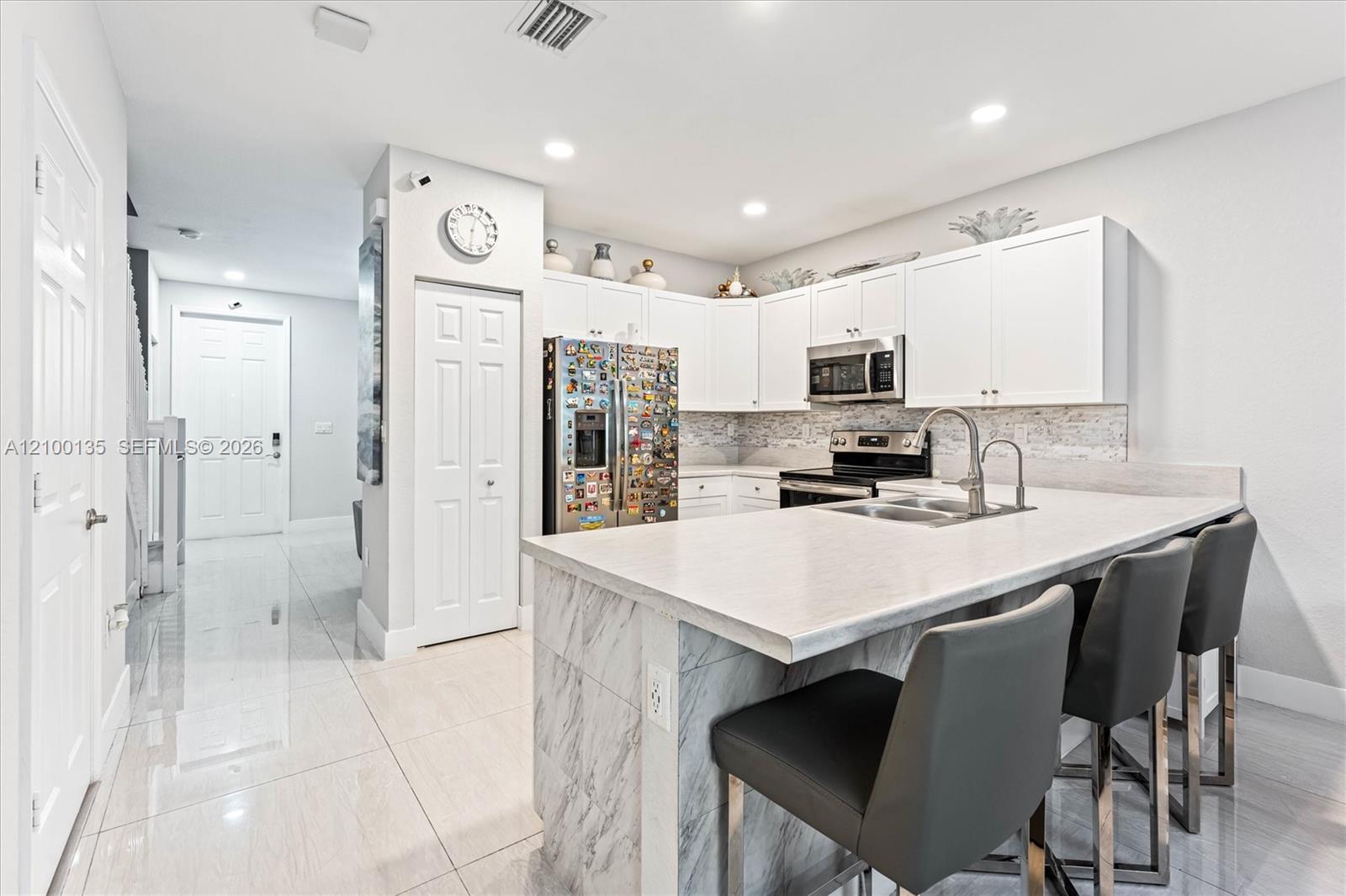 Kitchen with white cabinets, a marble-look island and backsplash, stainless steel appliances, and three gray bar stools.