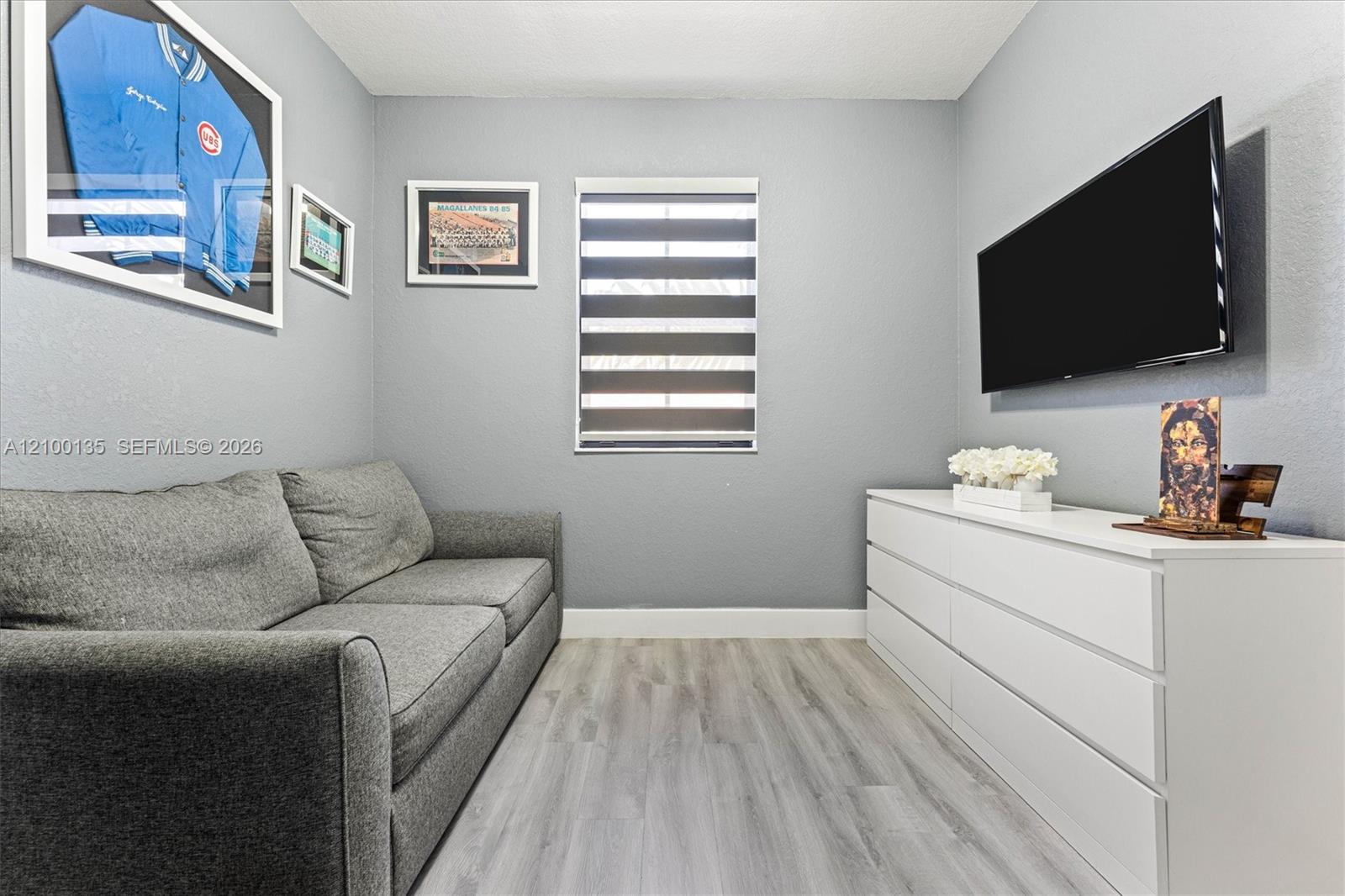 Furnished living room with a gray sofa, wall-mounted TV, white dresser, and framed baseball memorabilia.
