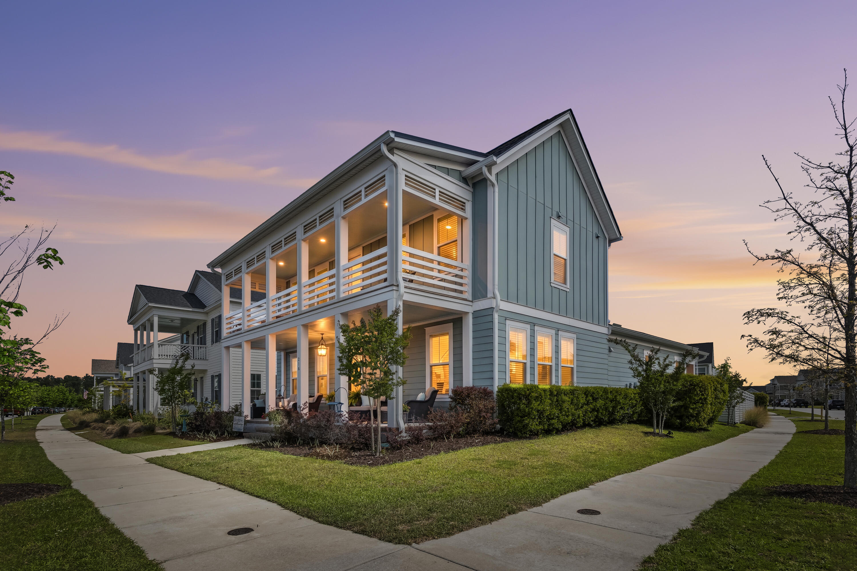 Two-story blue-gray home with a large white-railed porch glowing warmly at dusk.