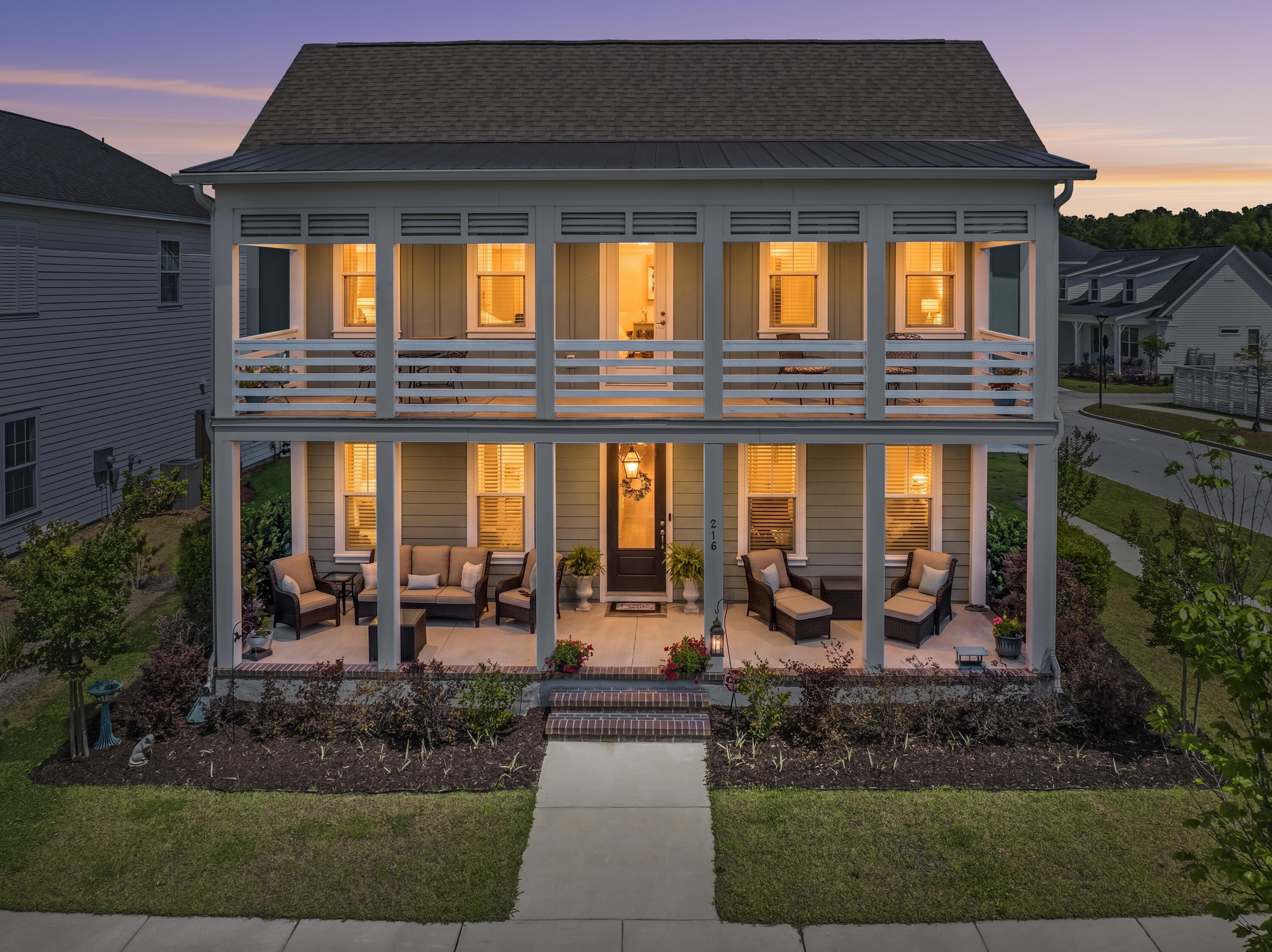 Two-story gray house with warmly lit, furnished porches on both levels at dusk.