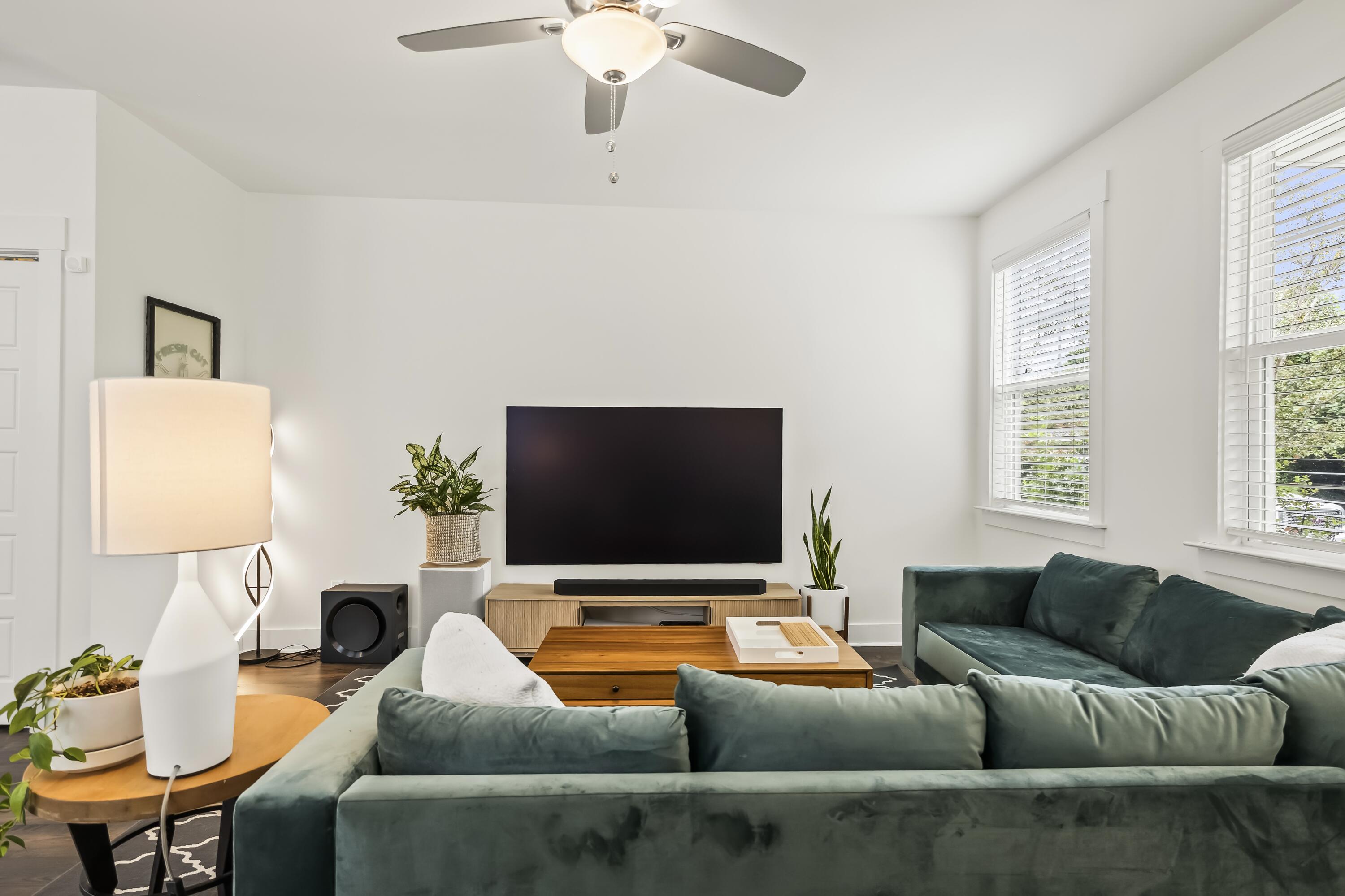 Living room with a dark green sectional facing a wall-mounted television, brightened by two windows.
