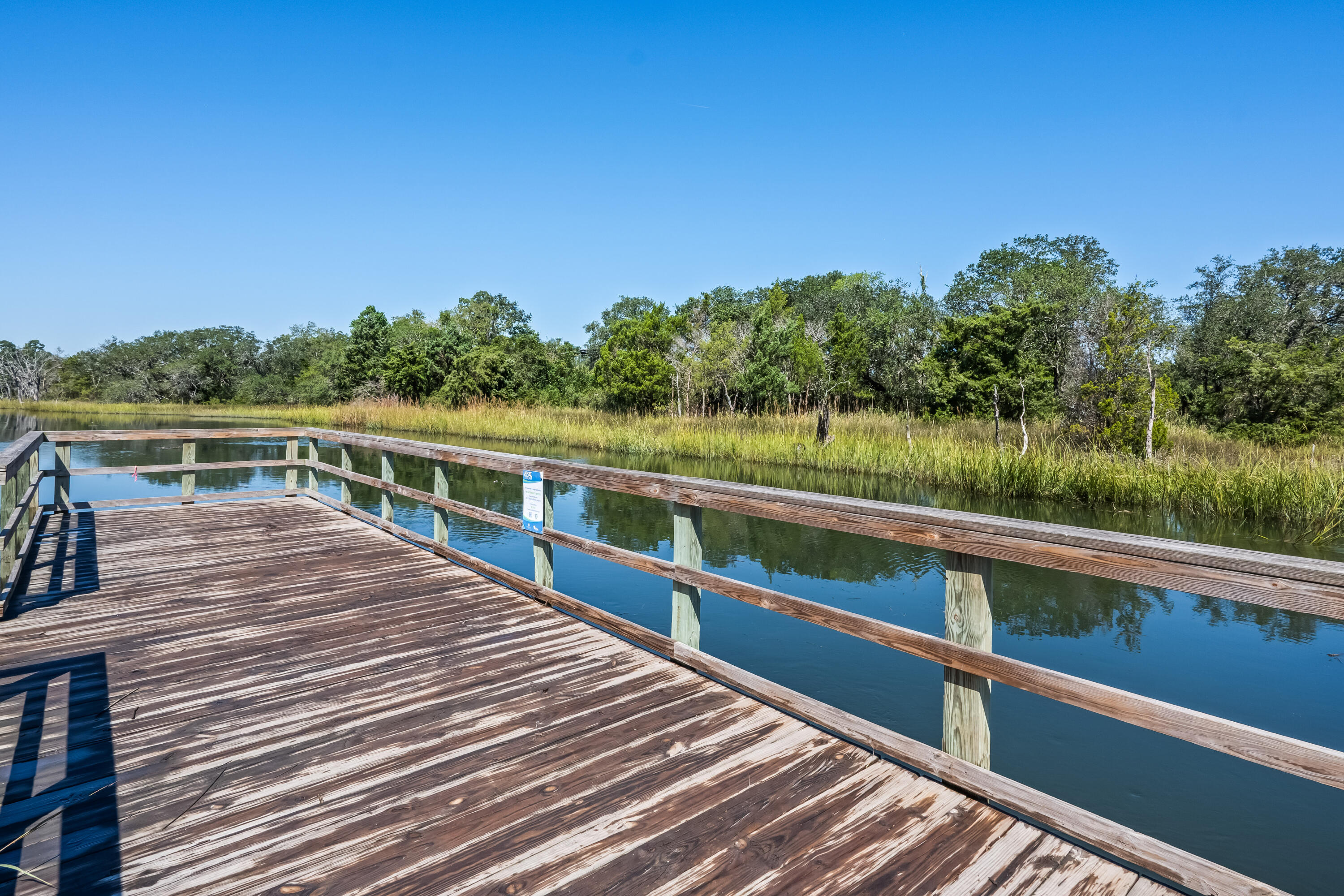 Wooden dock beside calm marsh water, with tall grasses and trees along the opposite bank.