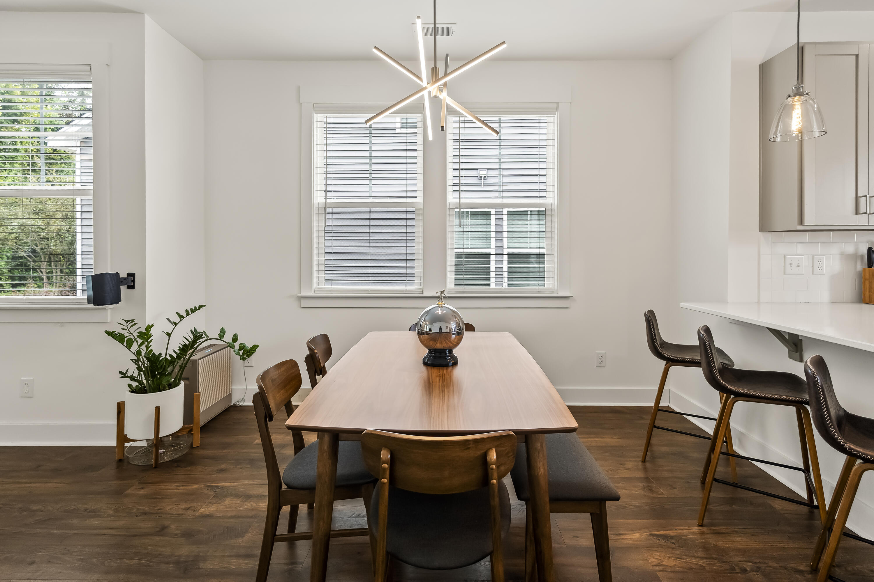 Dining area with a wooden table, modern chandelier, large windows, and kitchen counter with bar stools.