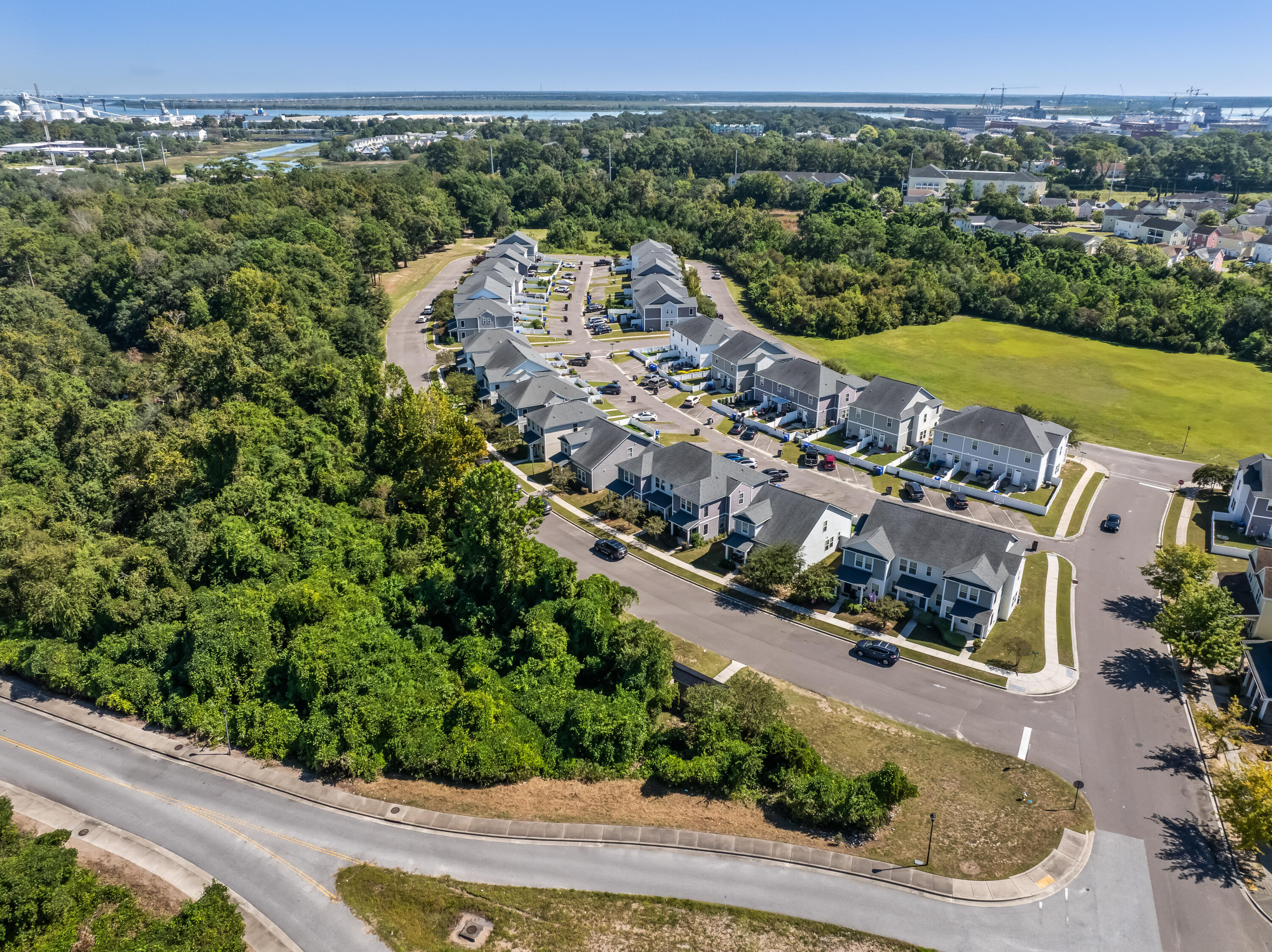 Aerial view of gray-roofed townhomes beside woodland and a grassy field, with water in the distance.