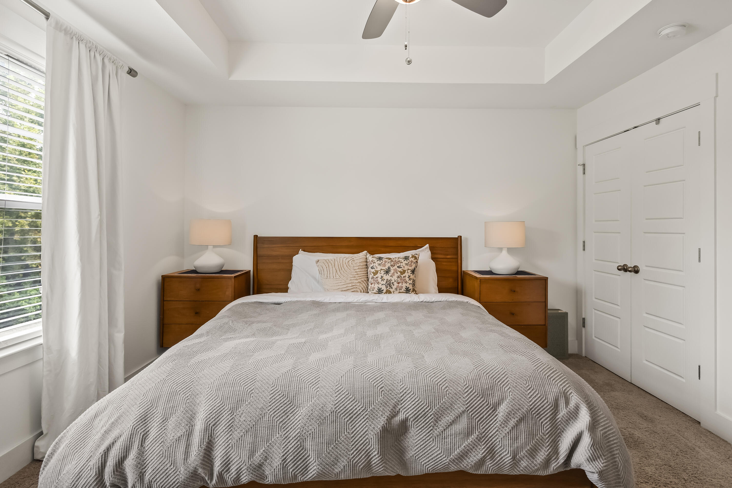 Bedroom with a white quilted bed, wooden headboard, matching nightstands, and curtained window.