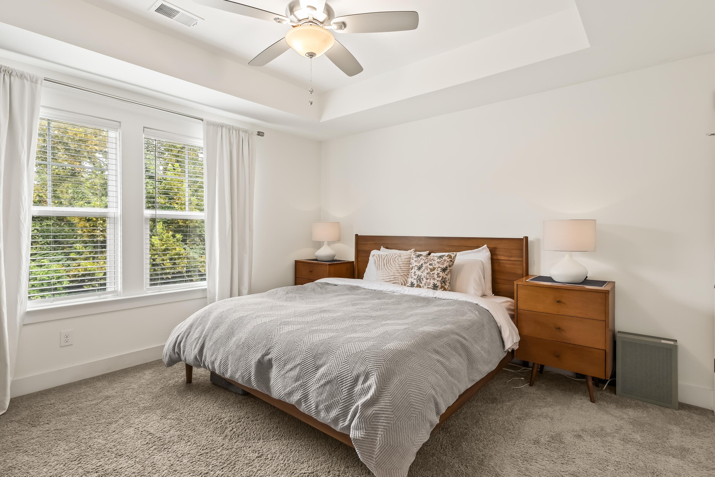 Bedroom with a wooden bed, gray patterned duvet, and two large windows overlooking trees.