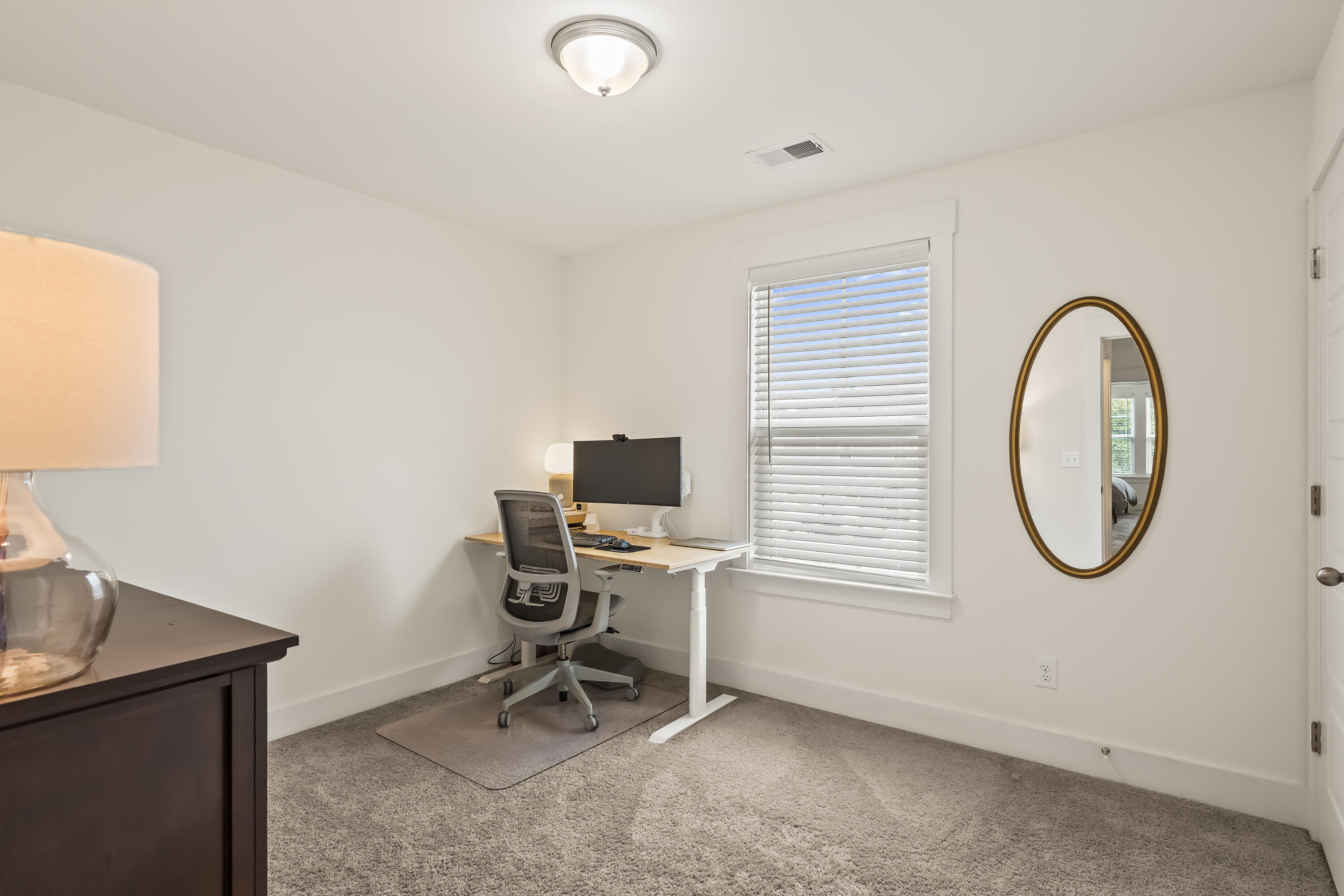 Home office with a standing desk and ergonomic chair beside a window, with an oval gold-framed mirror.