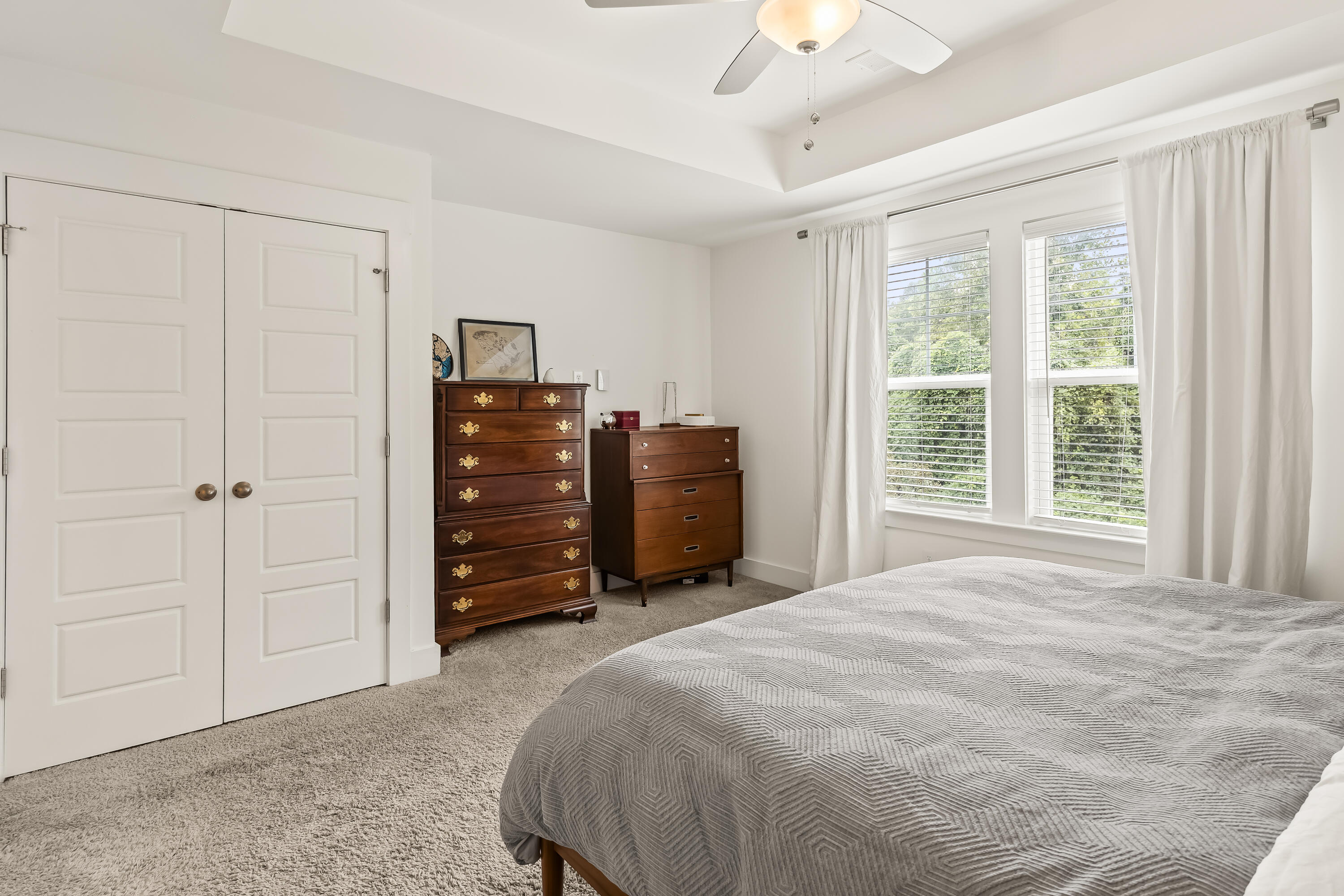 Bedroom with a gray quilted bed, wooden dressers, and large curtained windows.