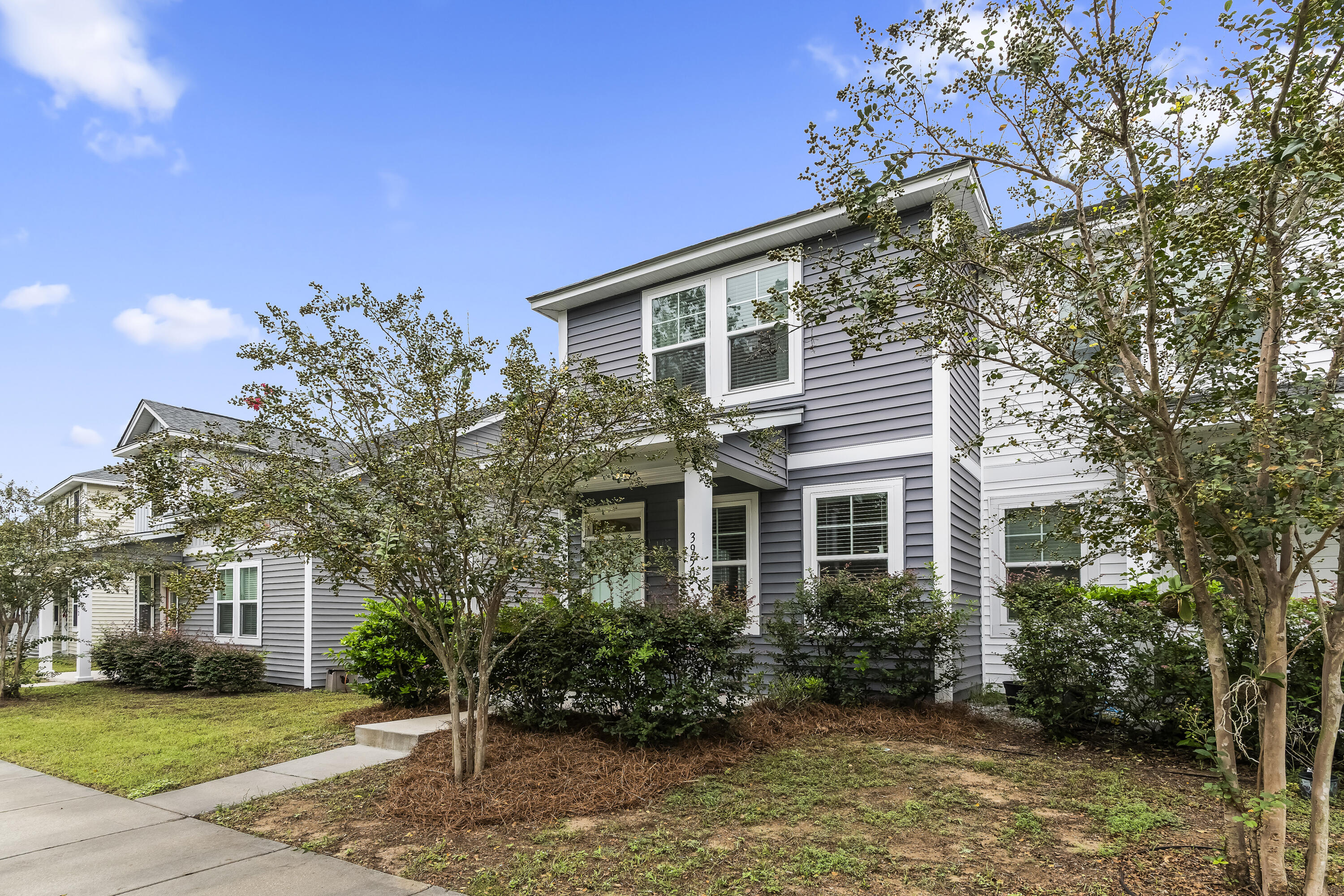 Two-story gray-sided house with white trim, framed by leafy trees and front-yard shrubs.