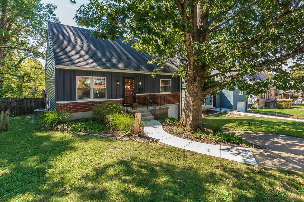 One-story dark-sided house with a brick facade, shaded by a large tree beside a curved walkway.