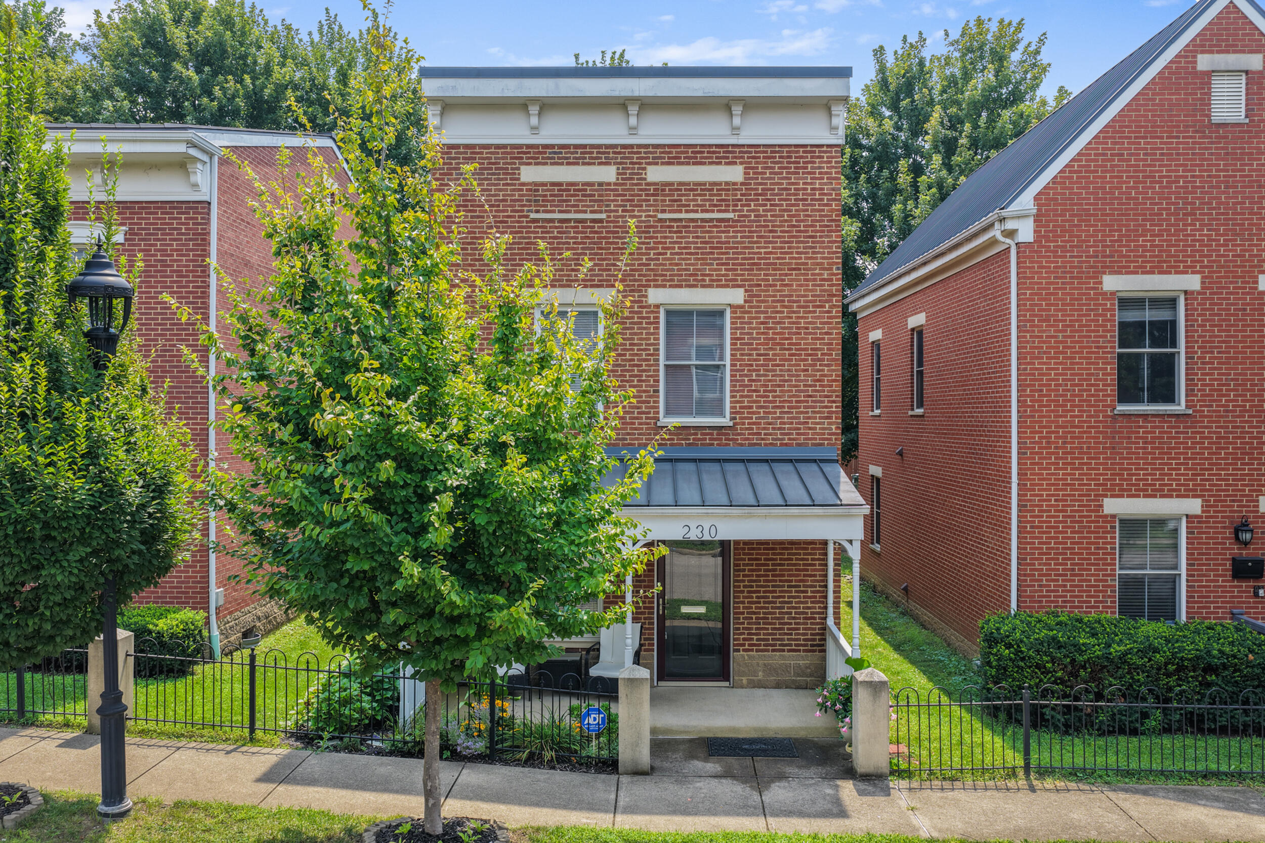 Red-brick residential building entrance marked 230, framed by leafy trees and fenced landscaping.