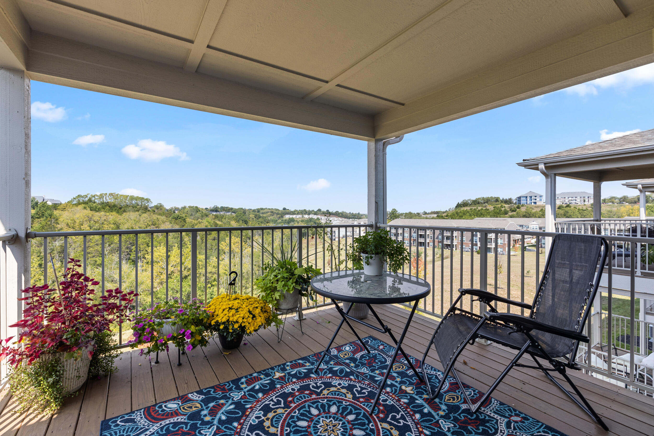 Covered balcony with patio seating, potted flowers, and a view of wooded hills and nearby apartments.
