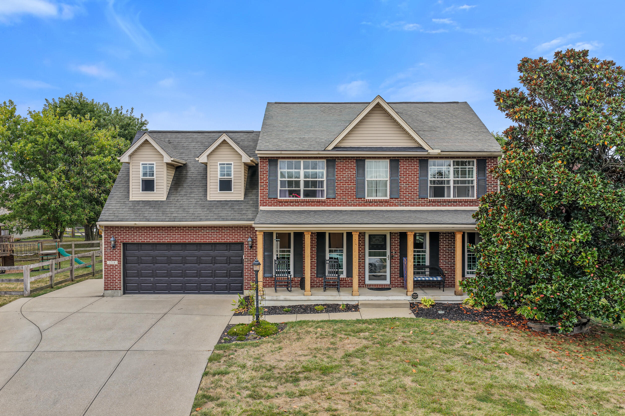 Two-story brick home with a covered porch, attached garage, wide driveway, and mature trees.