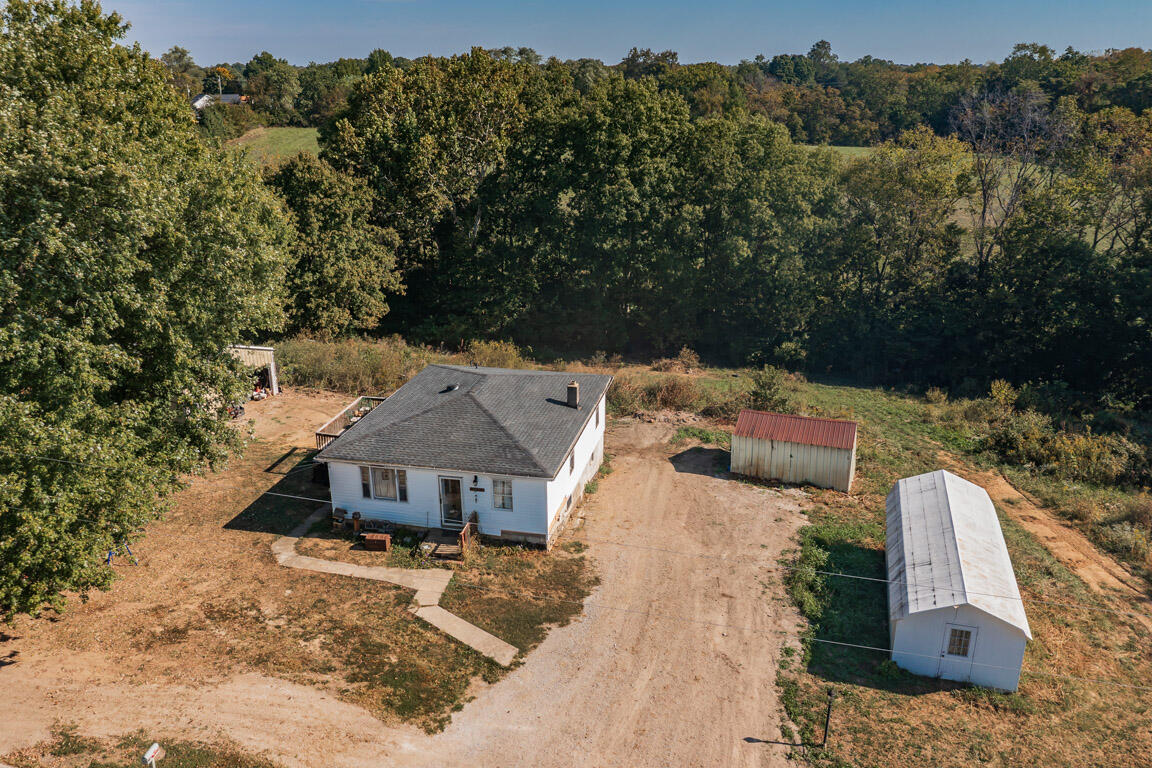 Aerial view of a white house, dirt driveway, and outbuildings on a wooded rural lot.