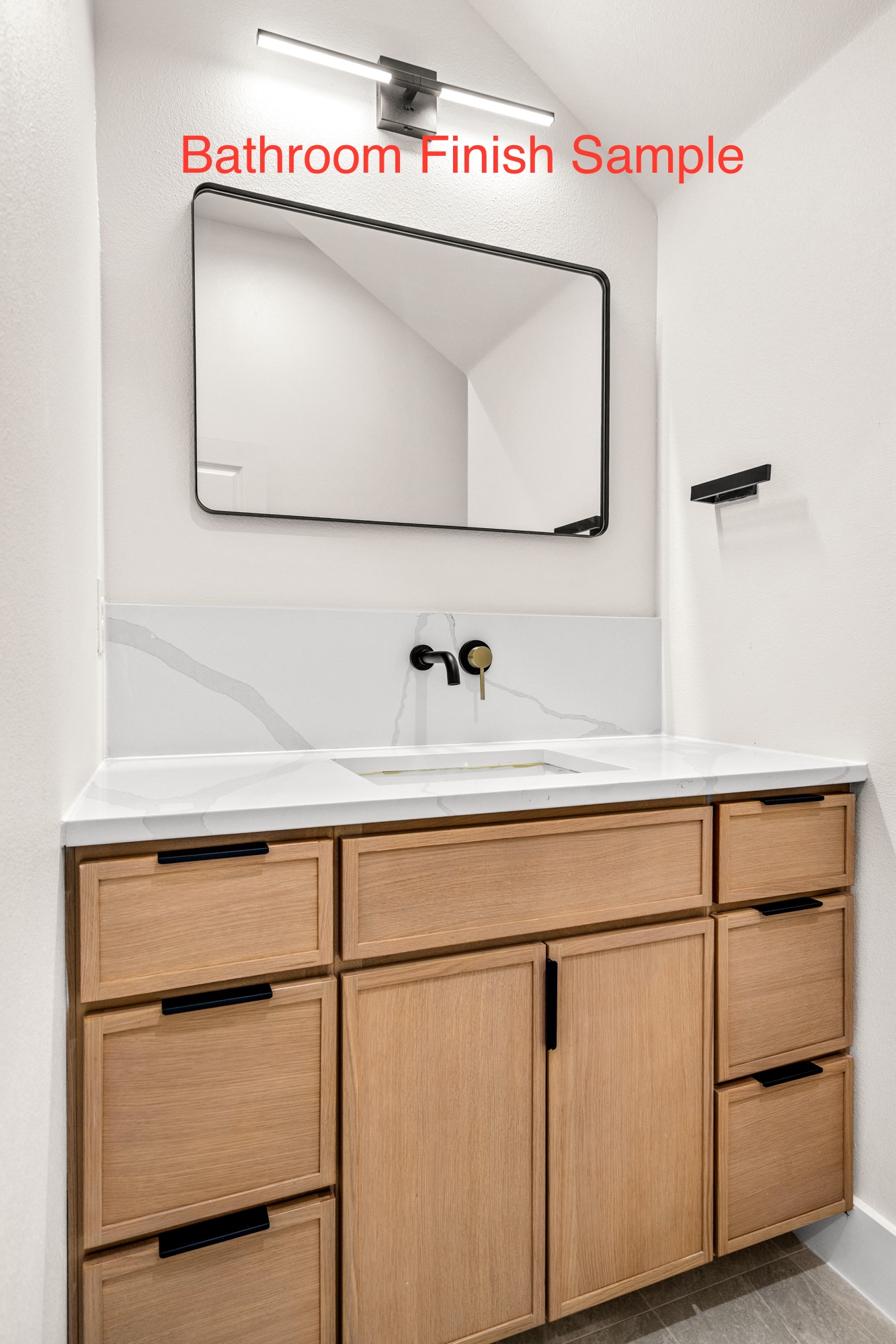 Bathroom vanity with light oak cabinets, veined white countertop, black-framed mirror, and black-and-brass fixtures.
