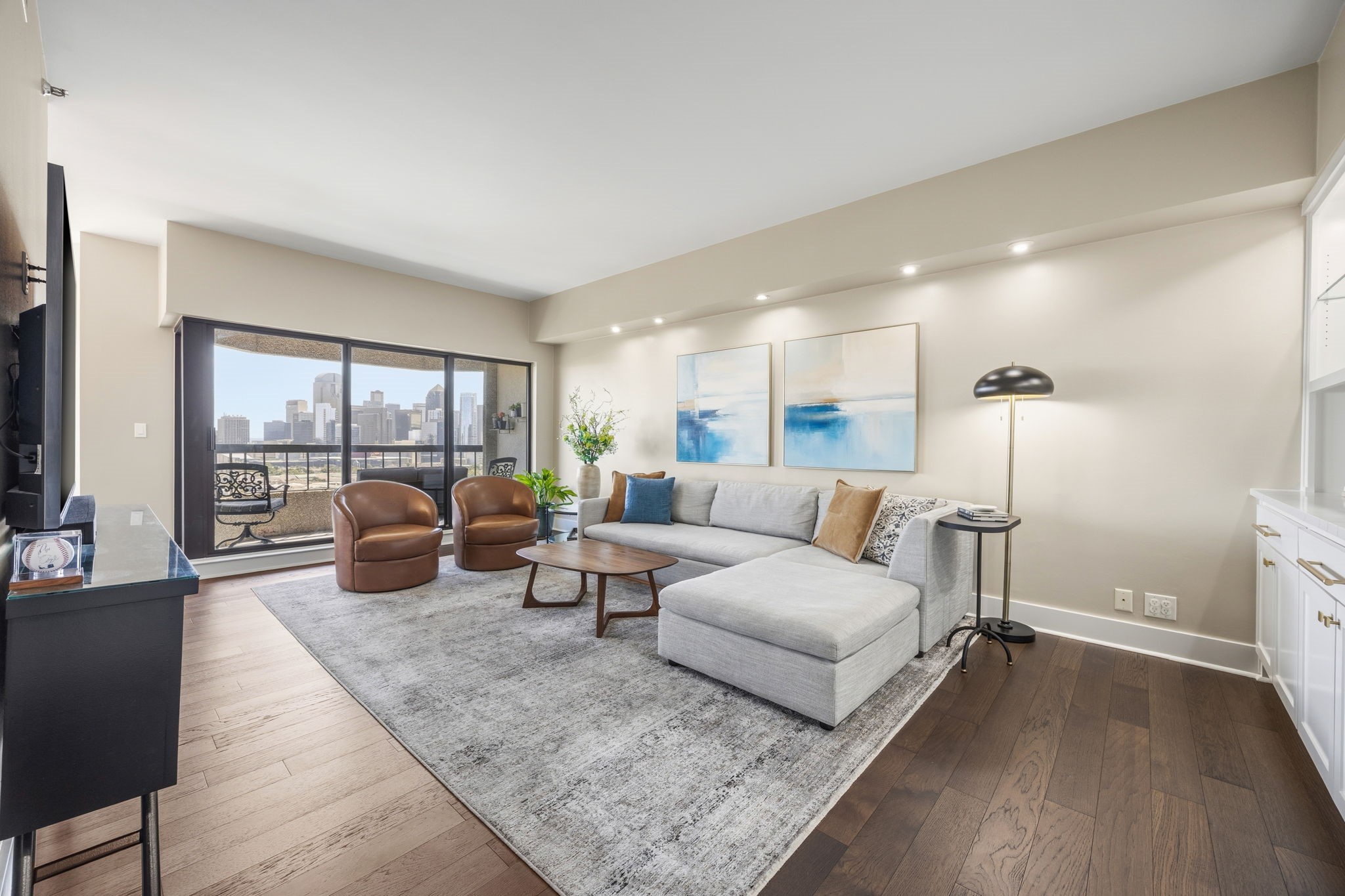 Staged living room with a gray sectional and balcony doors overlooking the Dallas skyline.