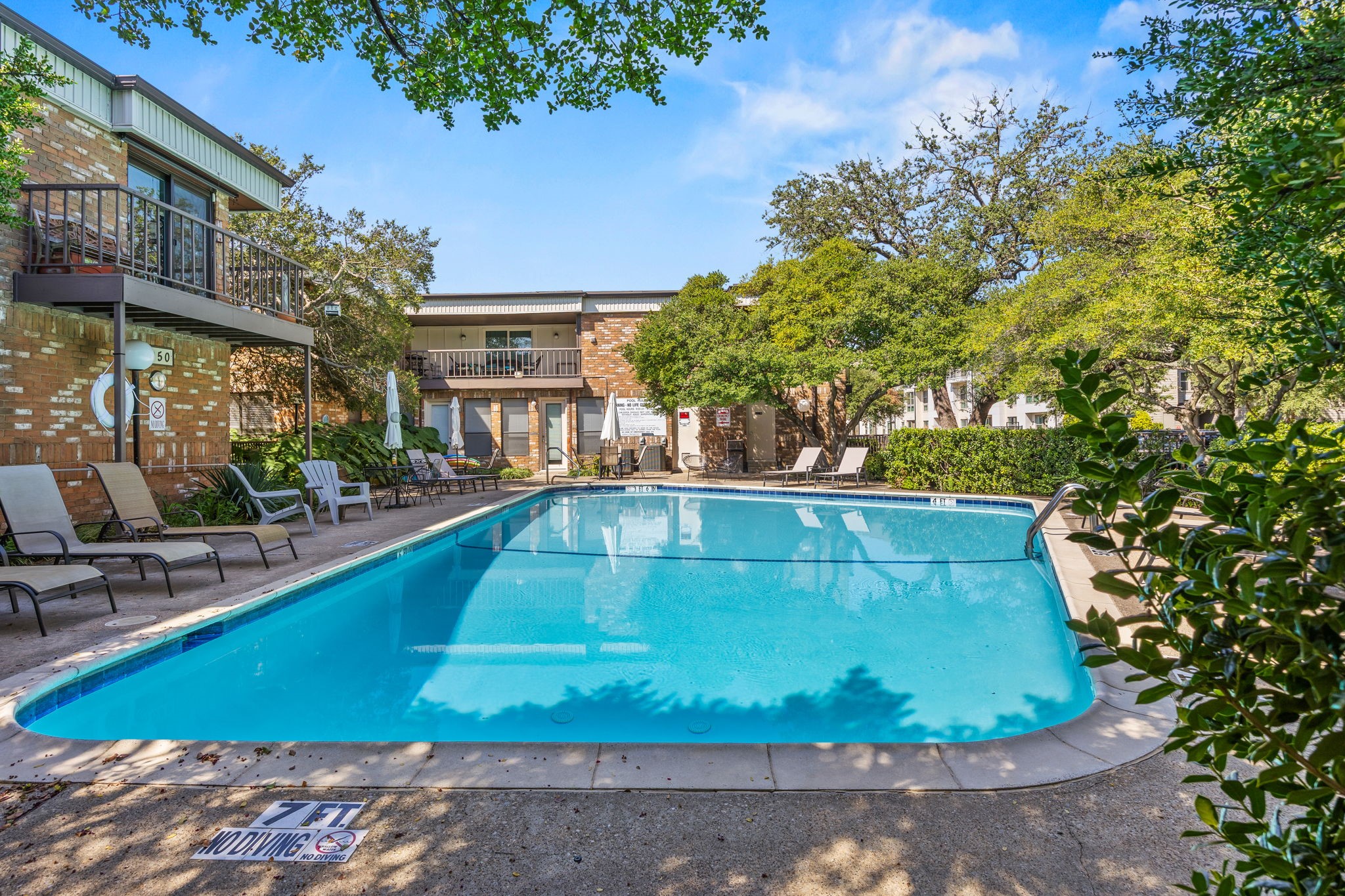 Outdoor pool at a brick apartment complex, with patio loungers and mature trees.